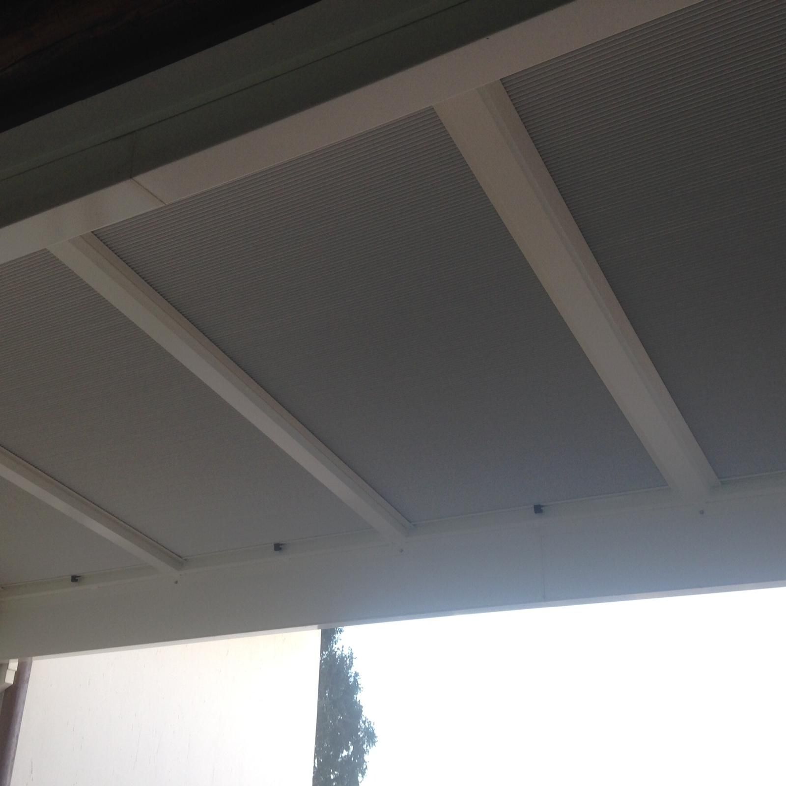 Vista dal basso di travi bianche a soffitto che incorniciano pannelli metallici grigi e perforati sul tetto di un patio esterno.