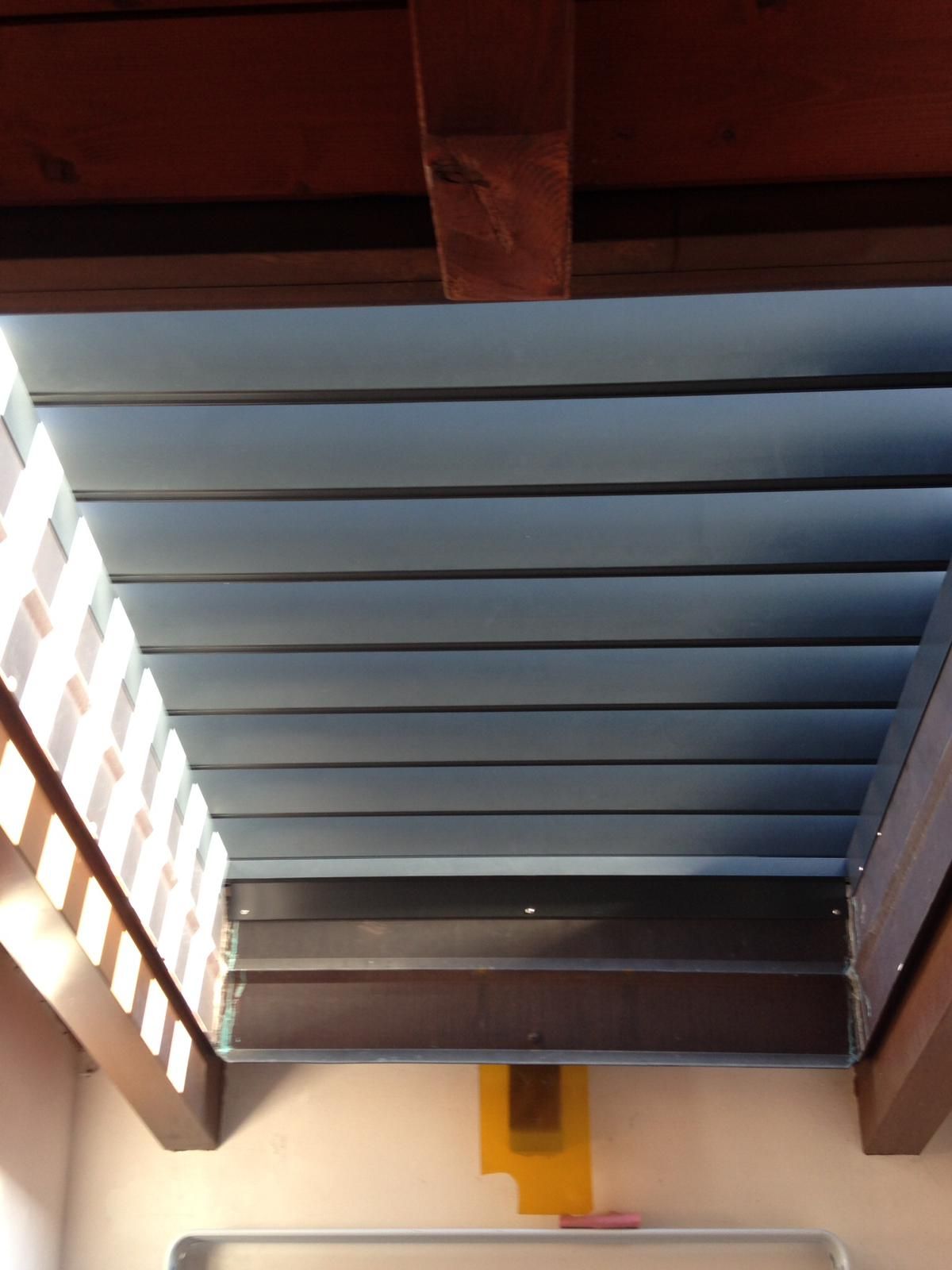 Una ripresa prospettica dal basso verso l'alto di una serie di tende orizzontali grigie incastonate tra due travi di legno scuro sul soffitto.