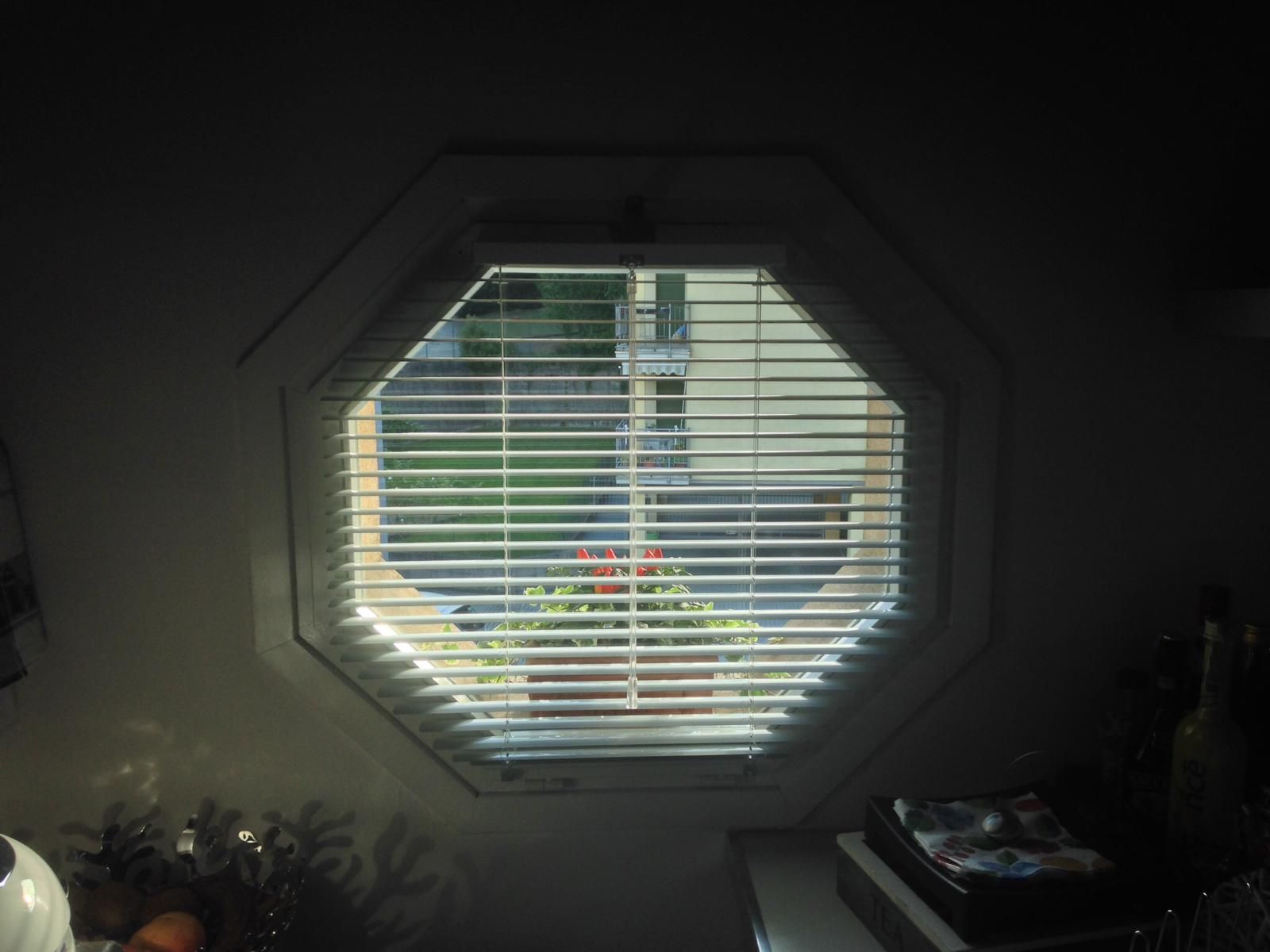 Una finestra ottagonale con persiane orizzontali chiuse che si affaccia su un prato verde e degli alberi.