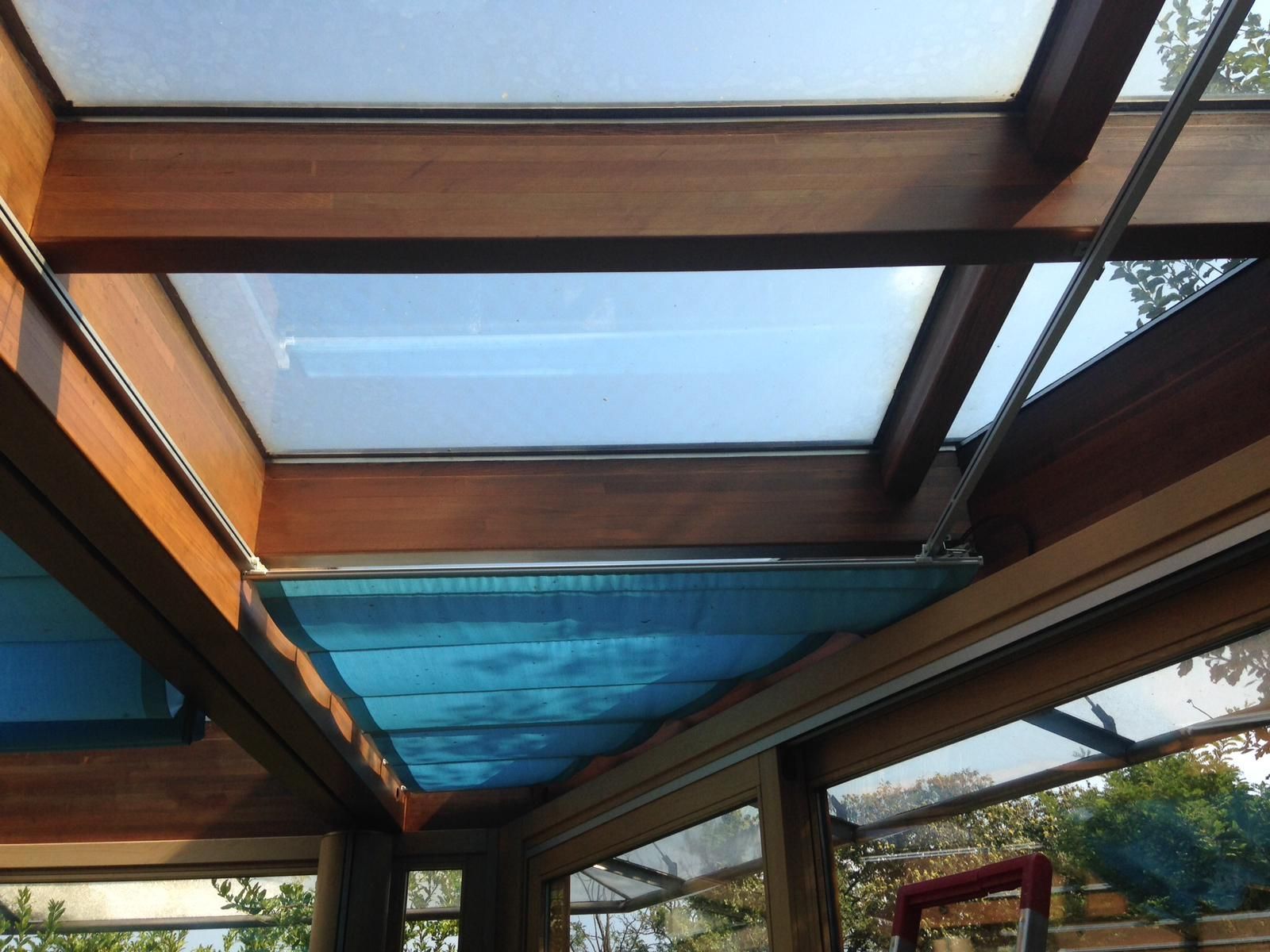 Soffitto di una veranda con struttura in legno, pannelli in vetro satinato e tenda a rullo in tessuto blu parzialmente estesa.