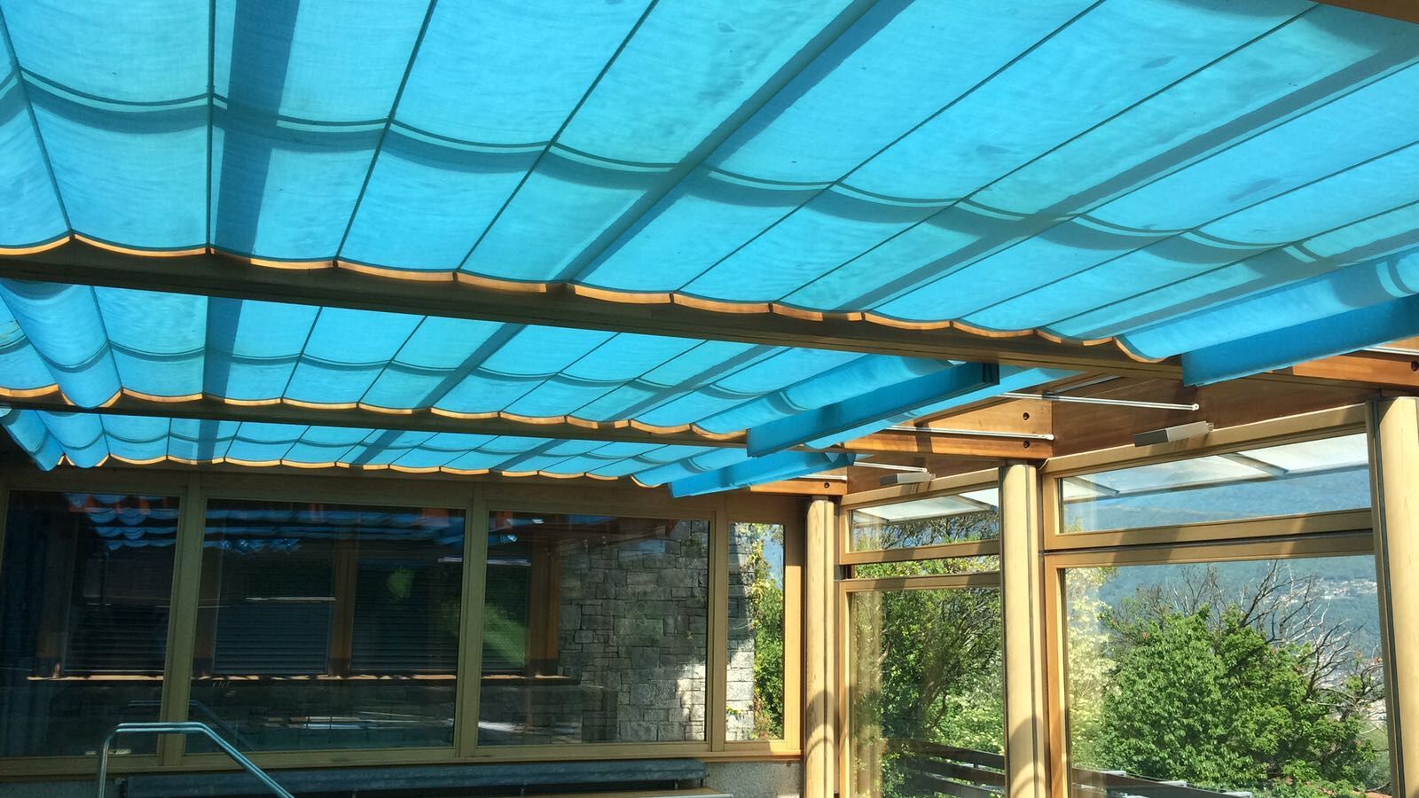 Una tenda parasole retrattile in tessuto blu brillante installata sotto il tetto di una pergola in legno, con vista sugli alberi e sul cielo.
