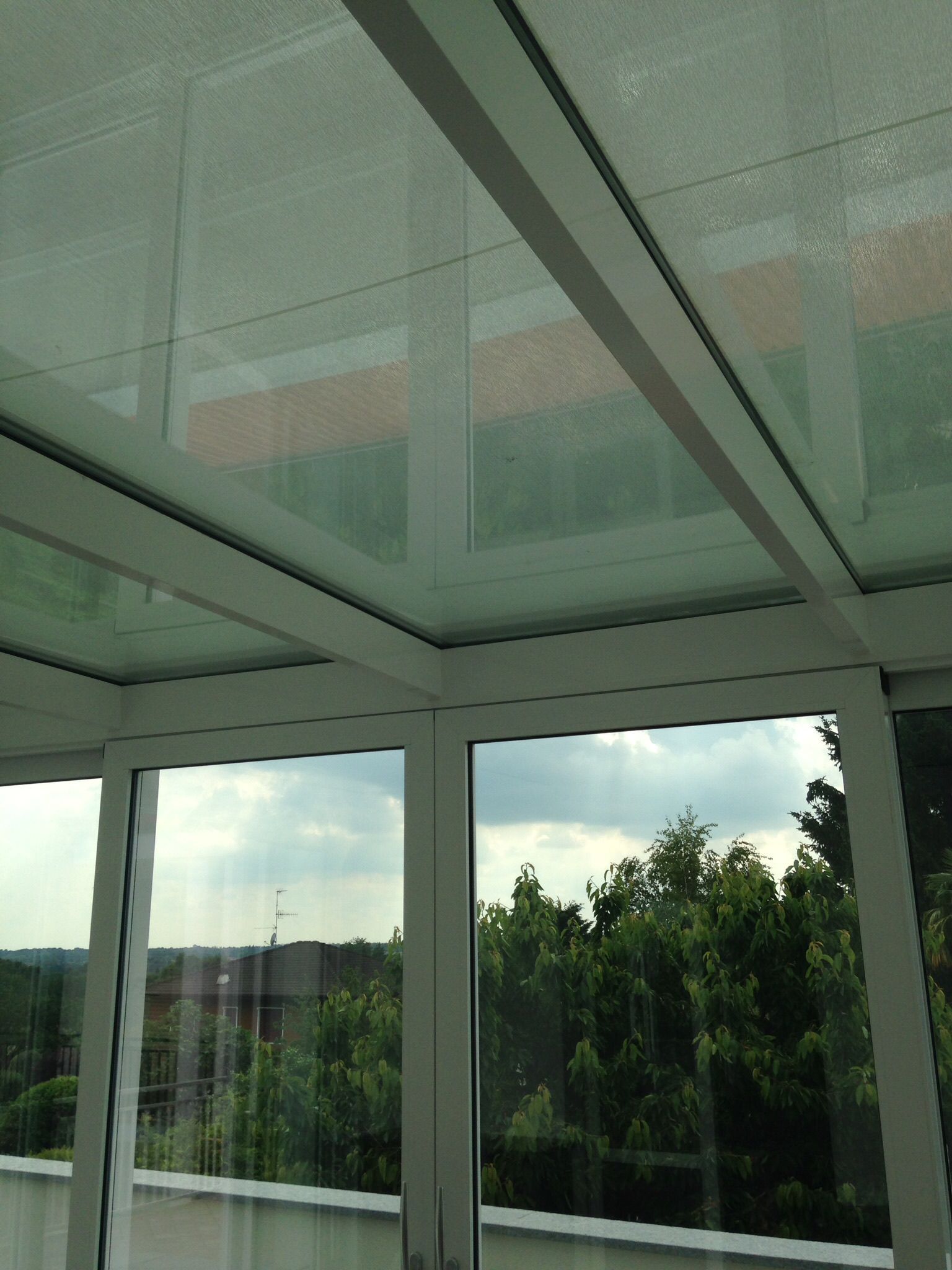Una vista dall'interno di una veranda che mostra un tetto in vetro con cornice bianca e finestre che si affacciano su alberi e un cielo nuvoloso.