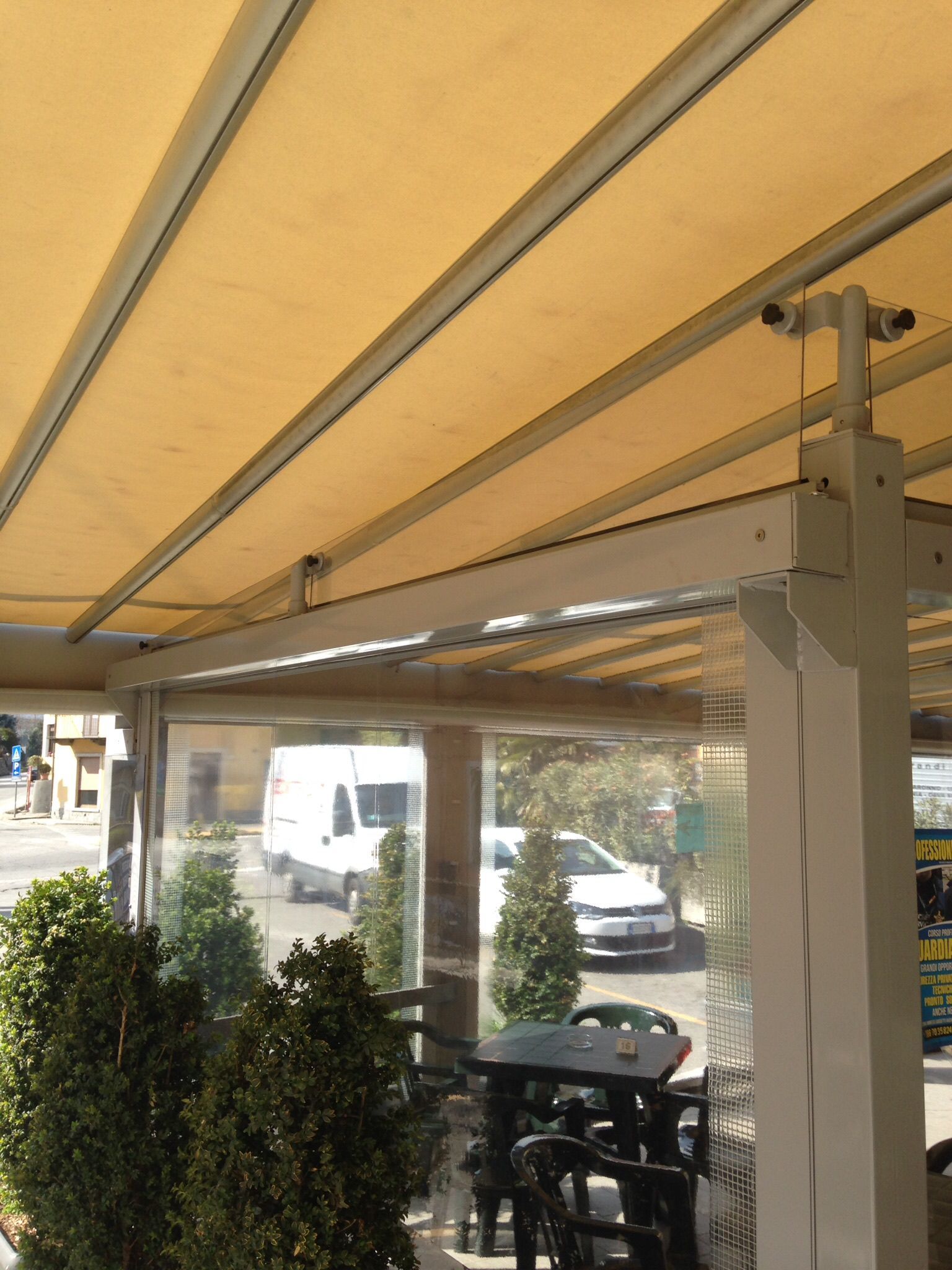 Area esterna con tavoli e sedie, coperta da una tettoia retrattile beige con tende laterali in plastica trasparente.