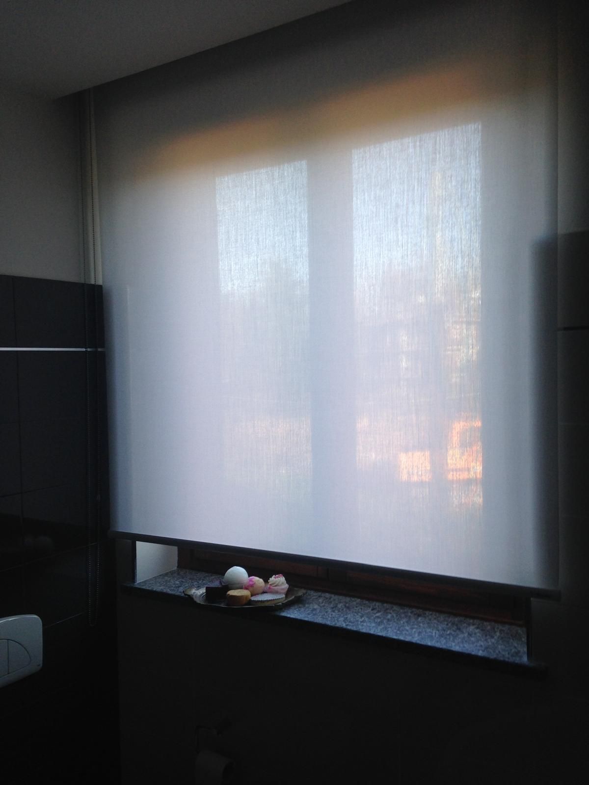 In una stanza dai toni scuri, una finestra con una tenda a rullo bianca traslucida sopra un davanzale in pietra, su cui sono appoggiati alcuni piccoli oggetti.