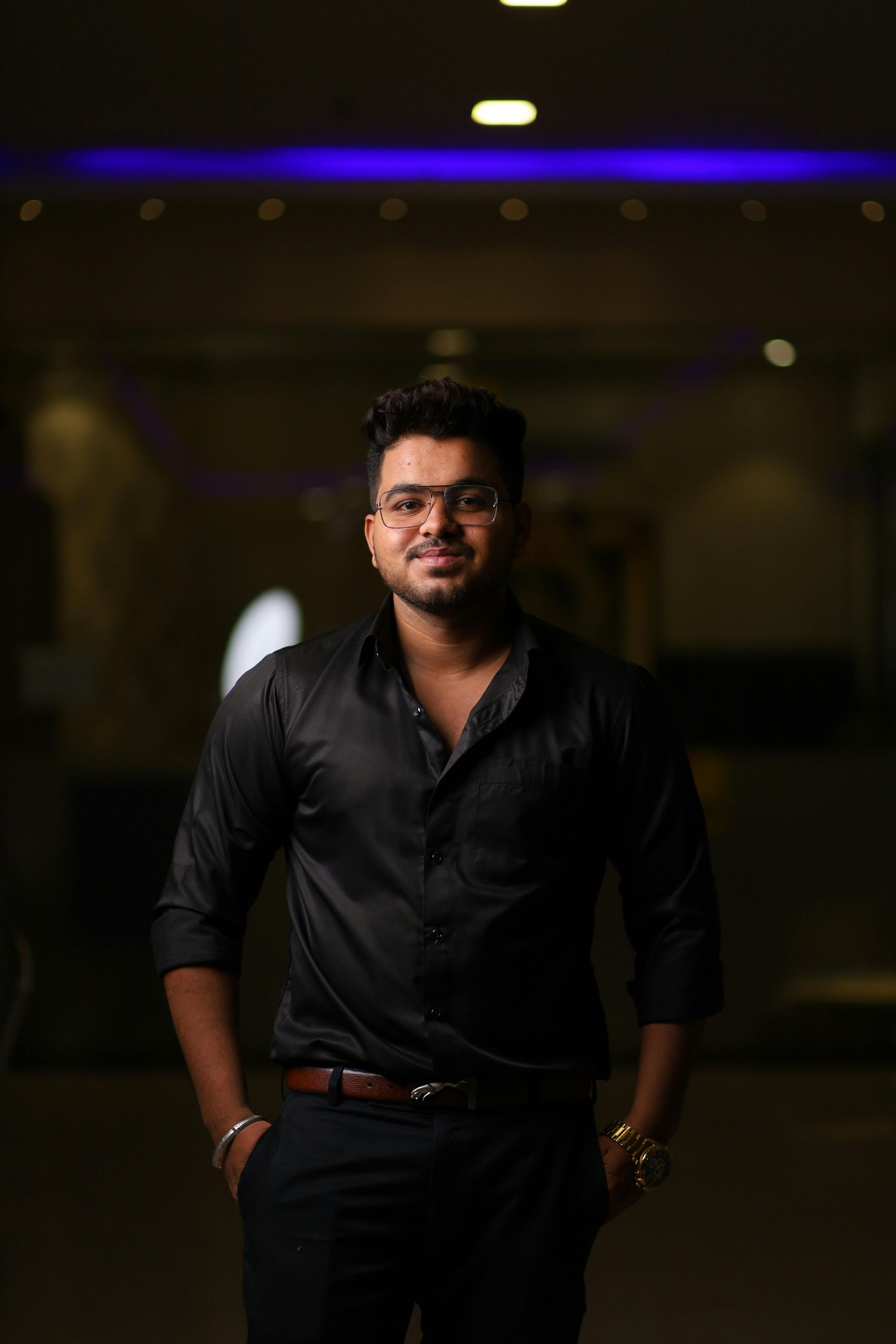 Man in a black shirt and trousers standing in a dimly lit hallway with blurred lights behind him