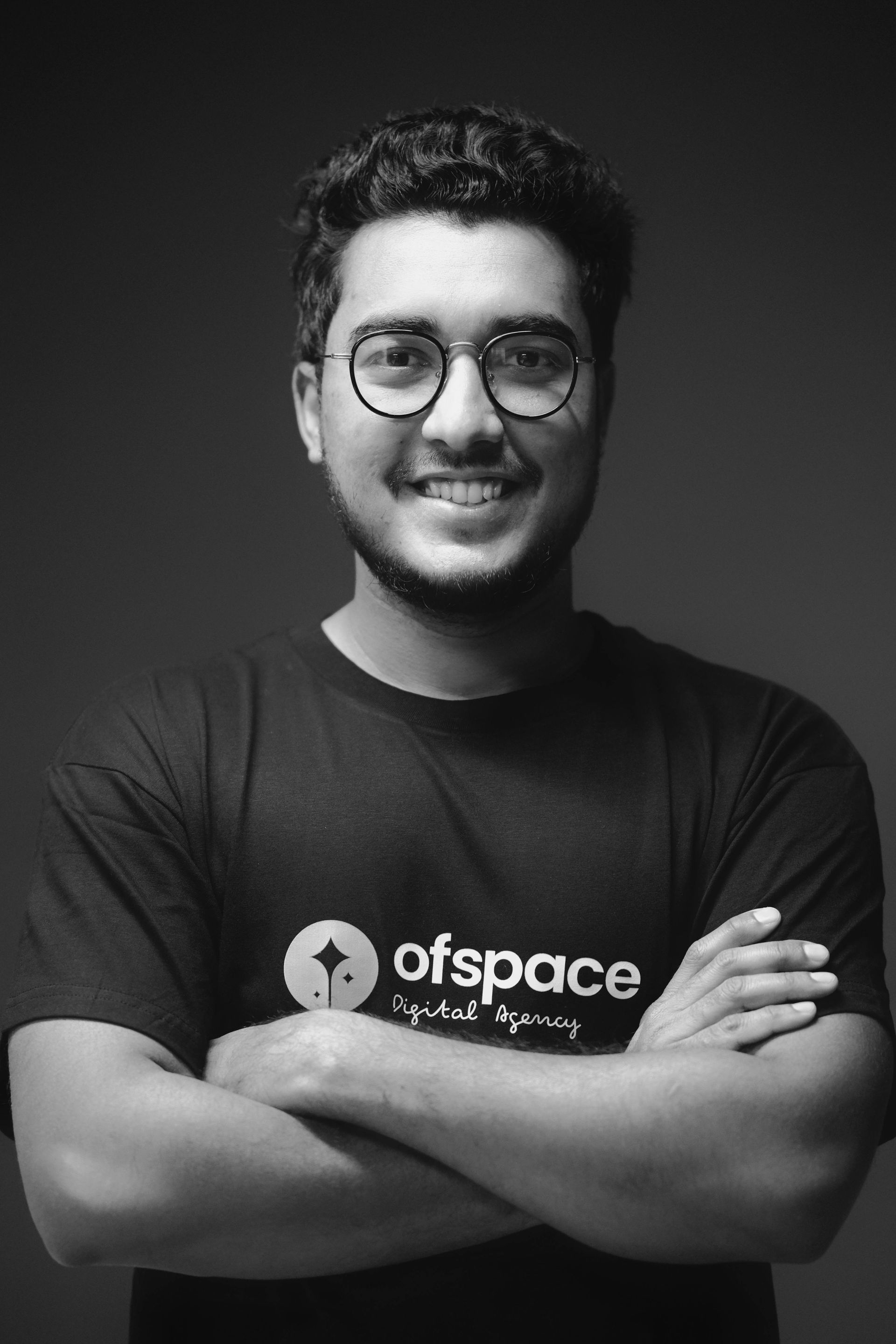 Smiling person with glasses, arms crossed, wearing a dark “ofspace” T-shirt against a dark background