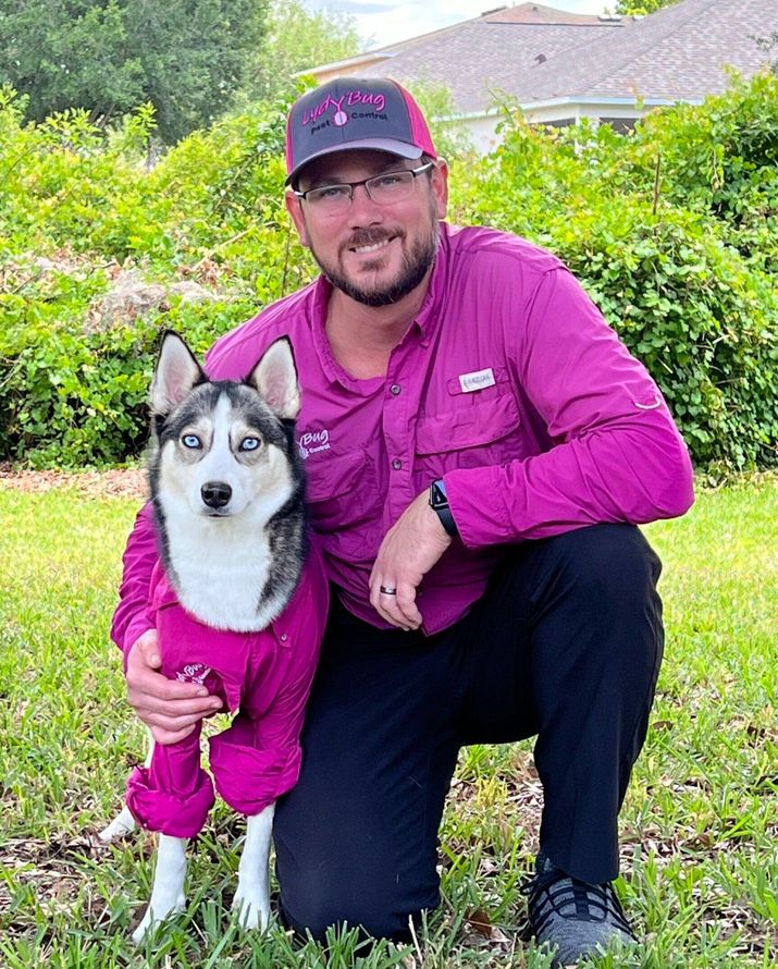 Man kneels beside a husky in a purple jacket in a grassy yard, both wearing matching purple