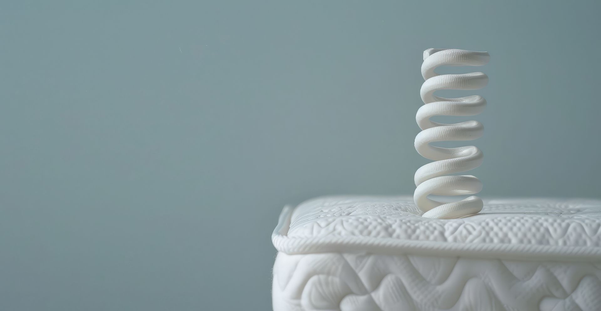 A white coil spring stands upright on the corner of a white mattress against a solid light blue background.