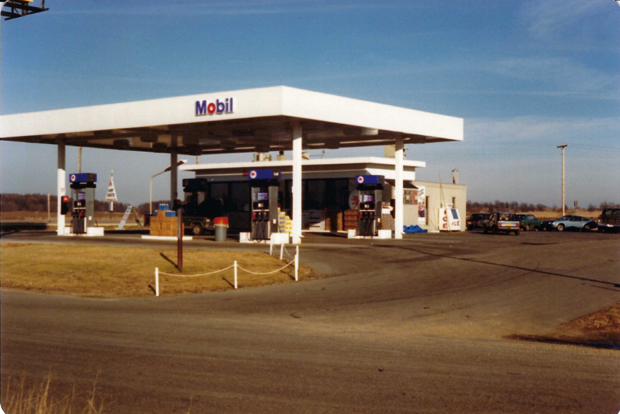 Mobil gas station with white canopy, pumps, and store on a sunny day. Cars parked nearby.
