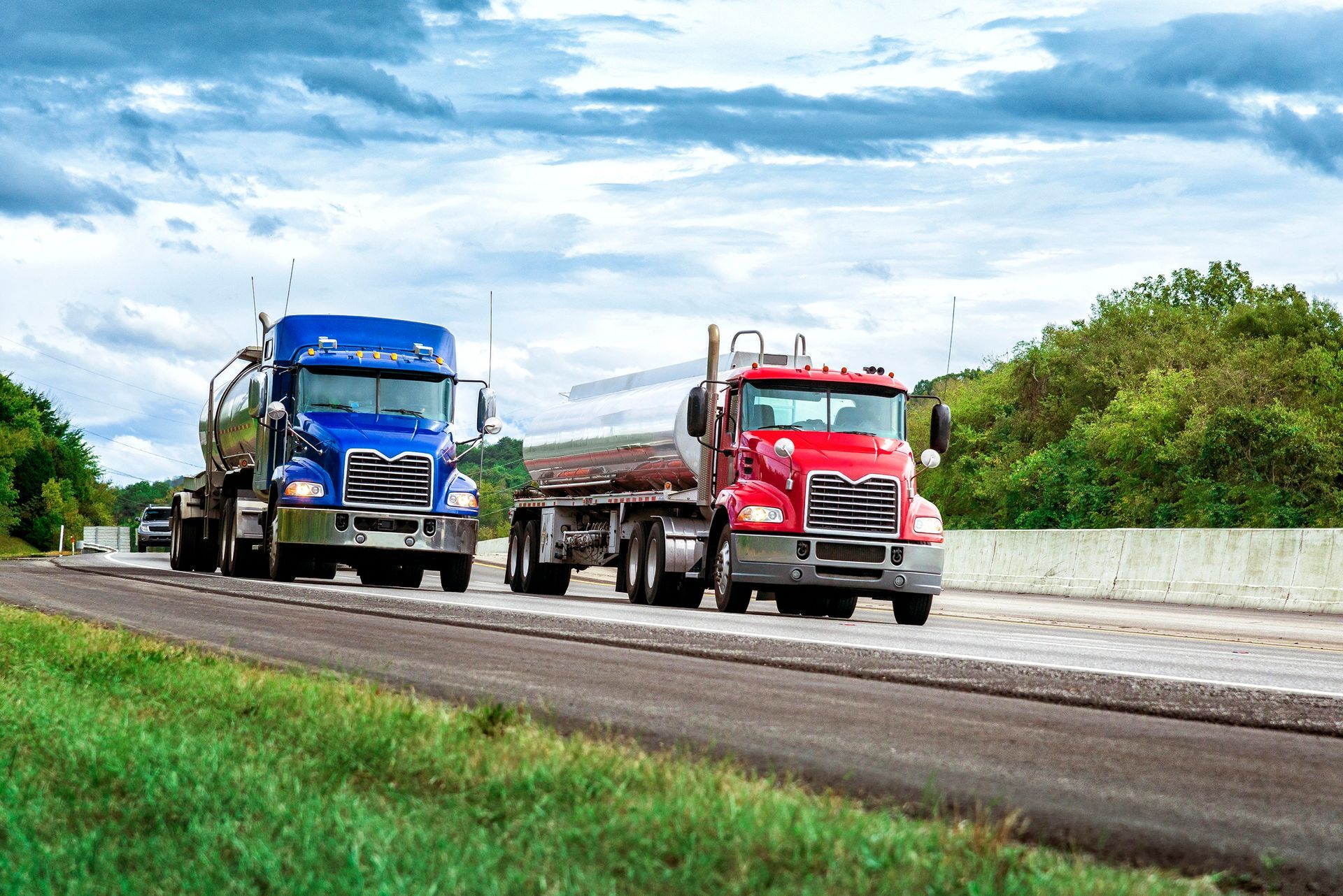 Two semi-trucks on a highway: a blue tanker and a red truck hauling logs, under a cloudy sky.