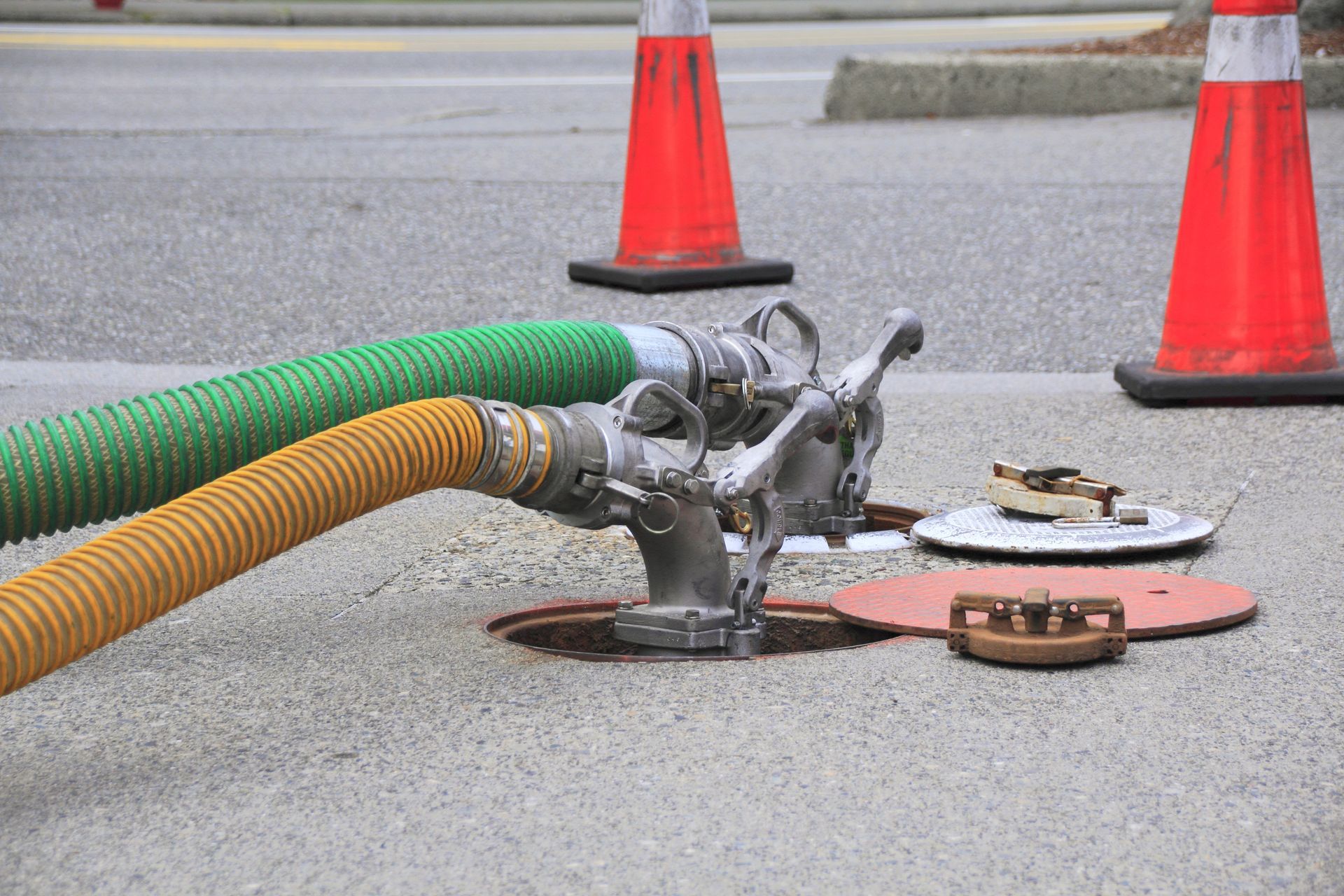 Hoses connected to an underground access point in a street, with orange traffic cones in the background.