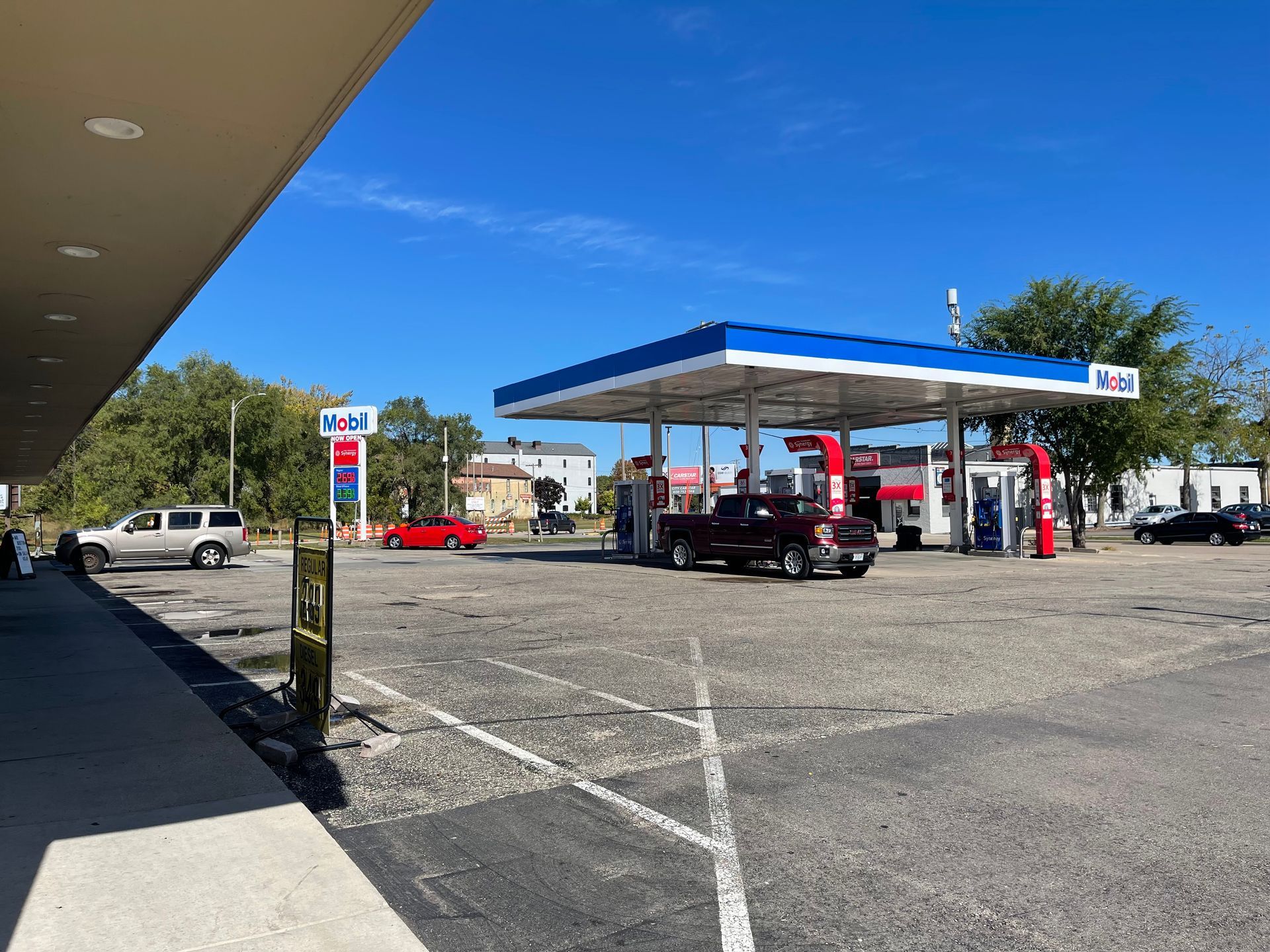 Gas station with a blue and white canopy. Cars at the pumps and a store front visible. Sunny day.