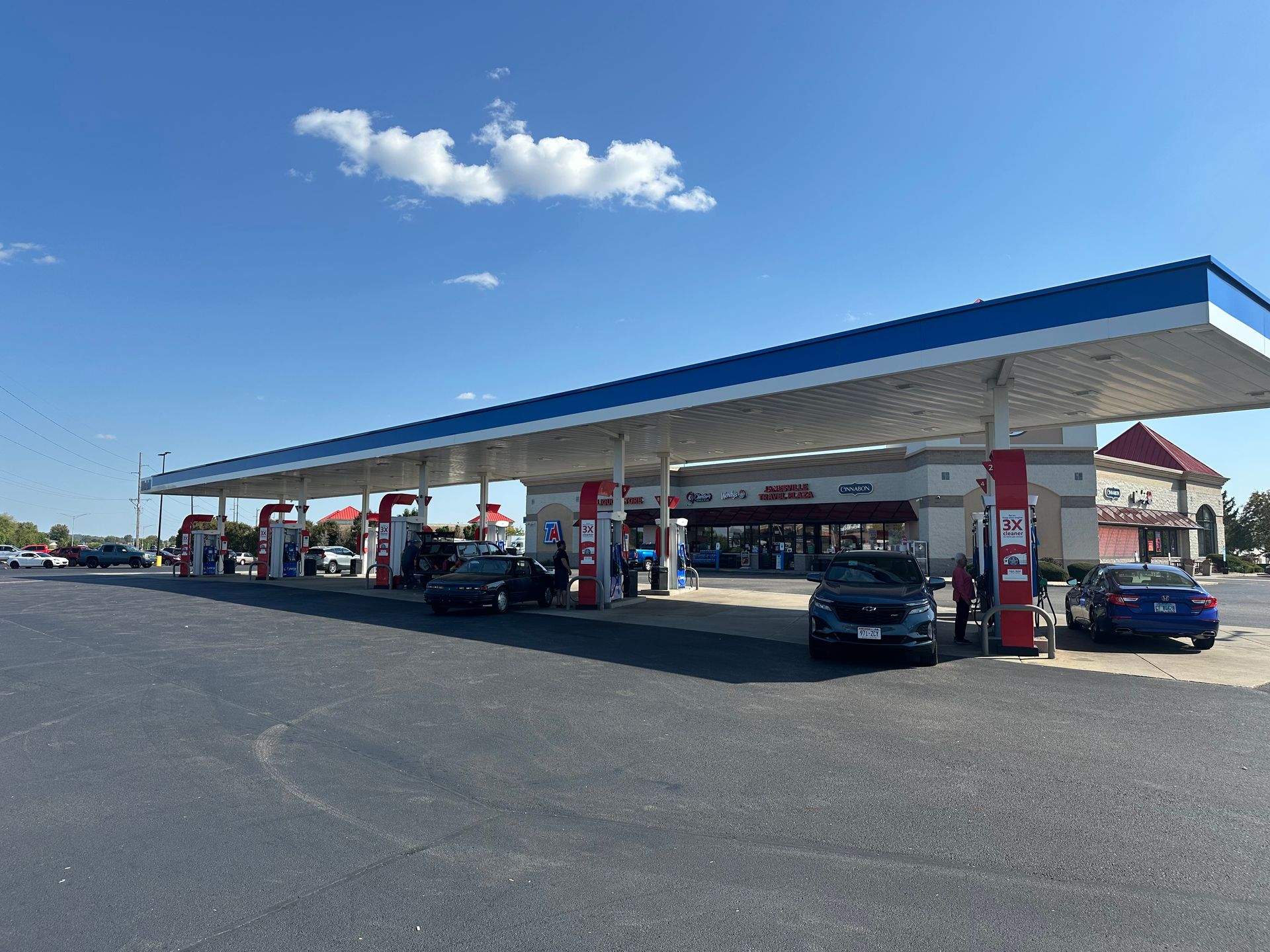 Gas station with cars at pumps, blue sky, white clouds, and store in the background.