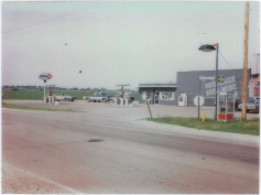 Gas station with blue signage, cars, and buildings under a cloudy sky.
