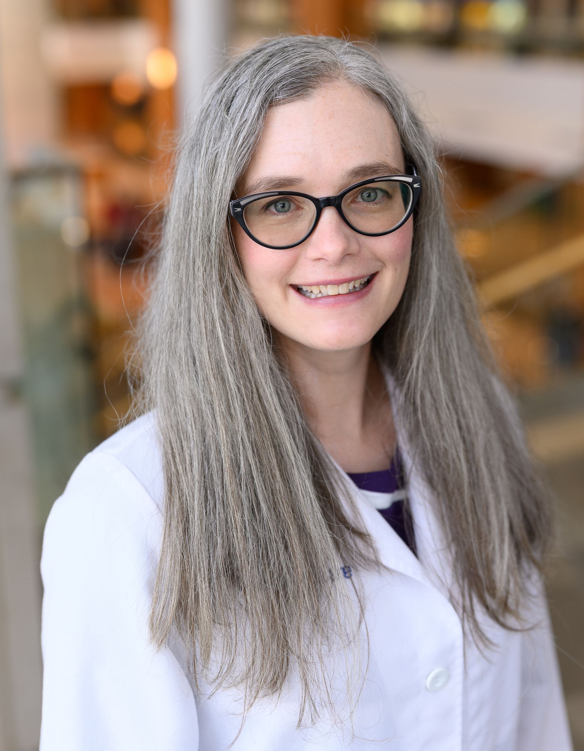 Woman with long gray hair, glasses, and white lab coat smiles, likely in a building.
