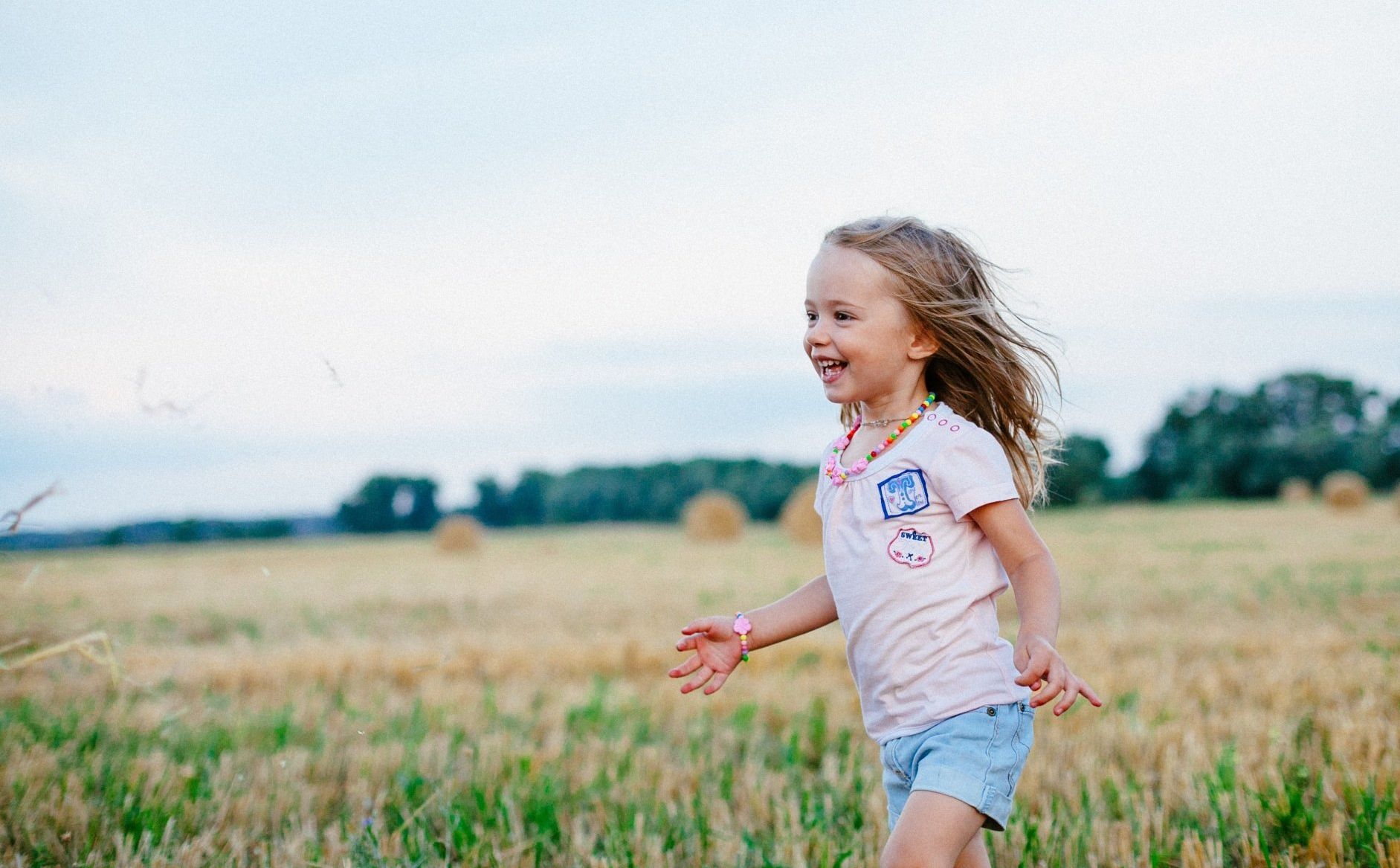 Girl running through a field, smiling. Wearing pink shirt and jean shorts, with hay bales in the background.