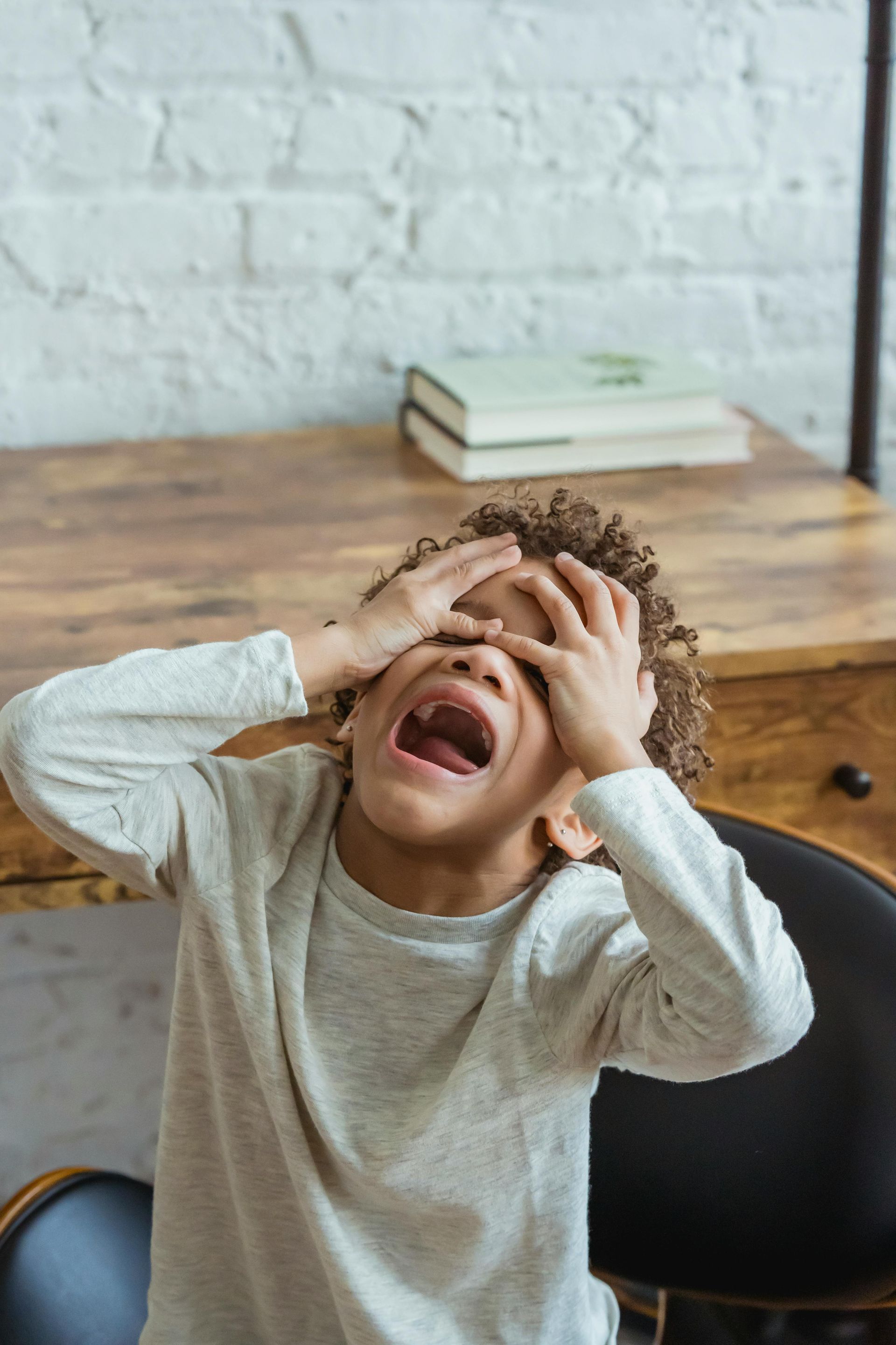Young child with hands covering eyes, screaming in distress near a desk with books.
