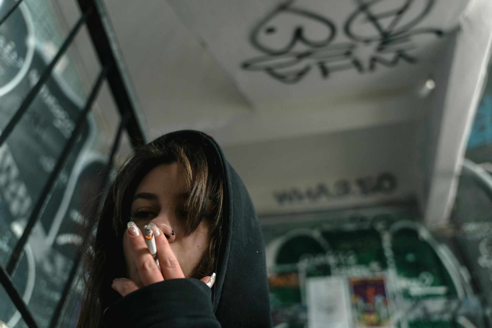 Woman in a black hoodie smoking a cigarette in a graffiti-covered stairwell.