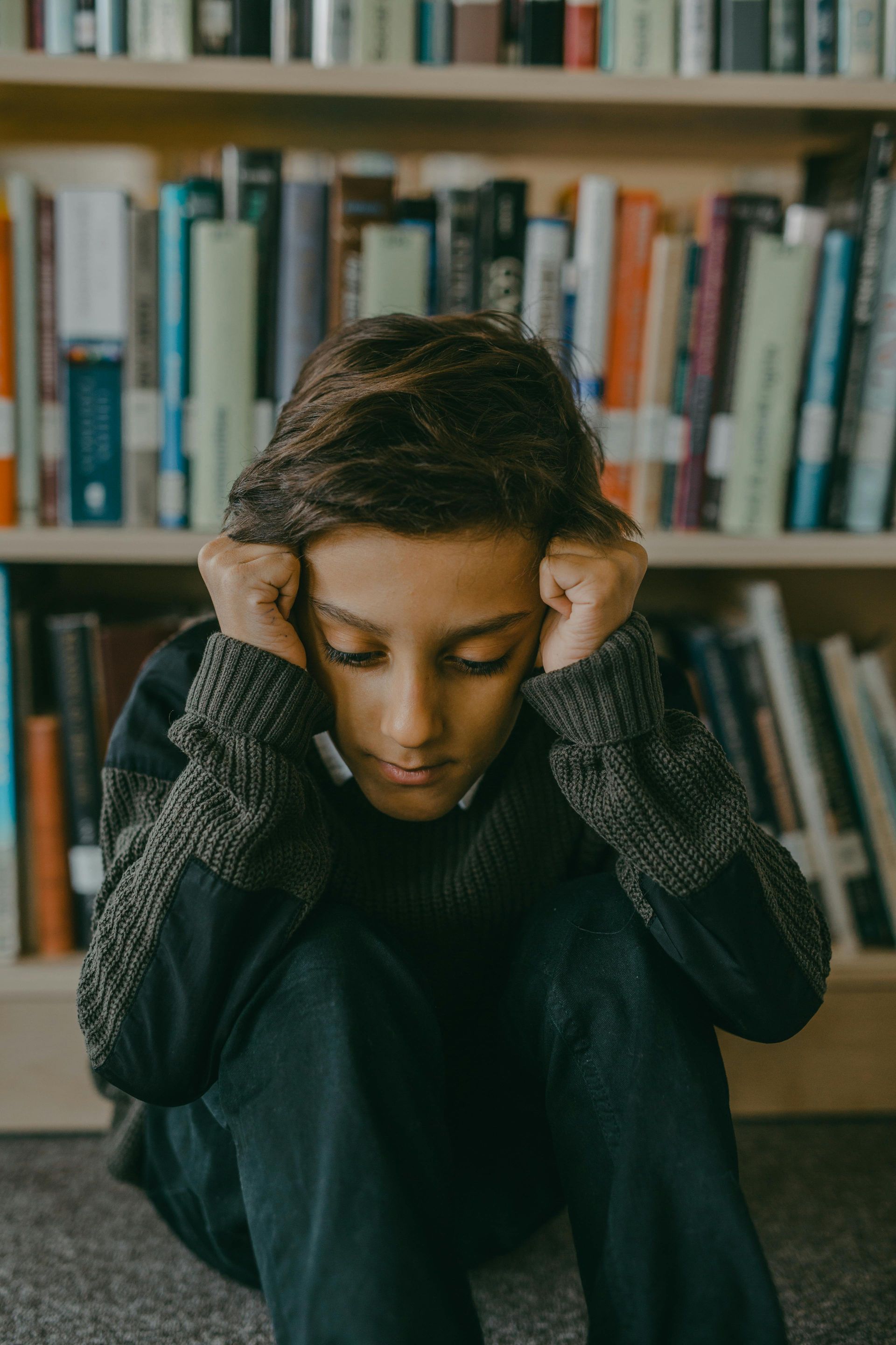 Boy with head in hands, sitting in front of a bookshelf, looking down, possibly stressed.