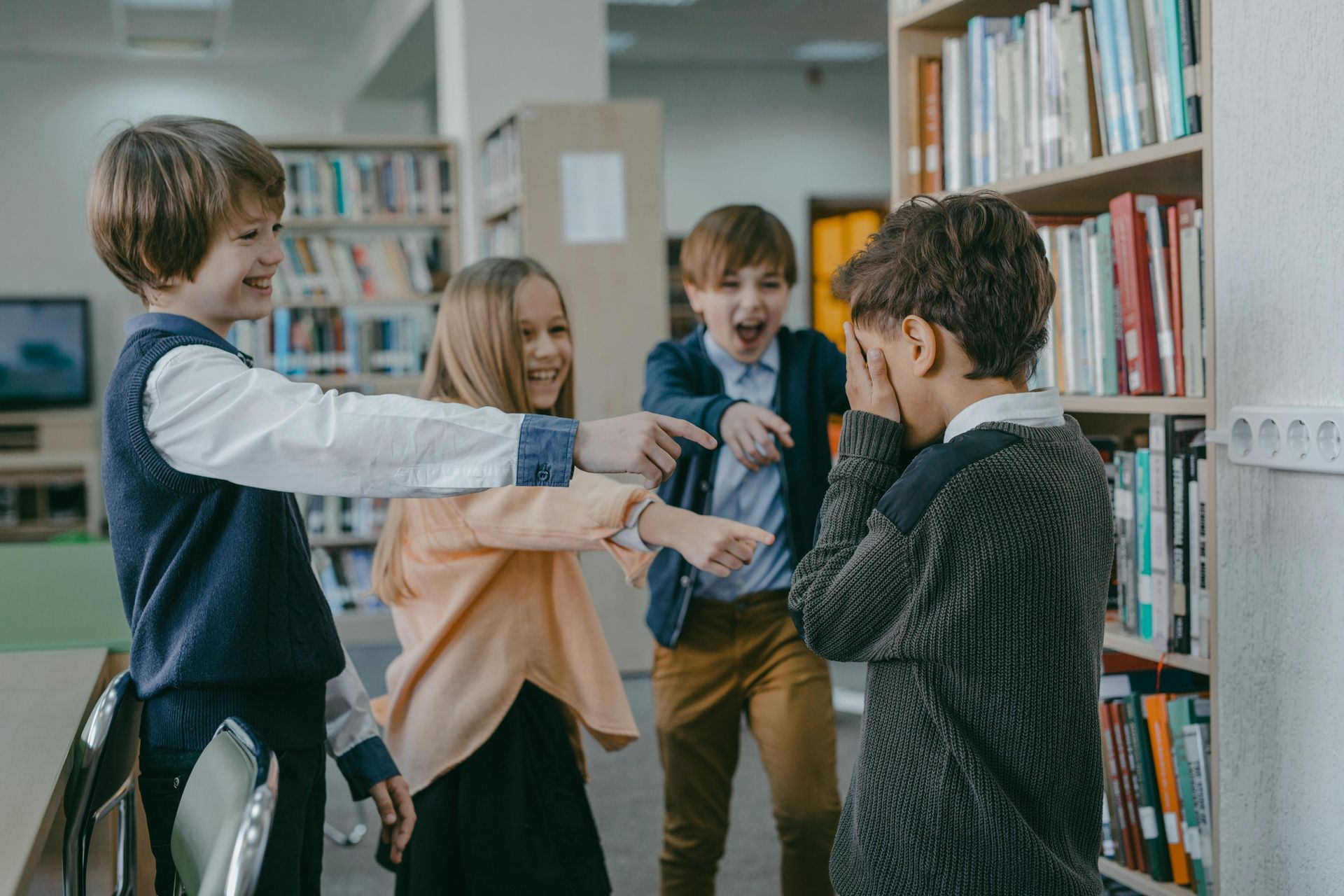 Four children in a library; three are laughing and pointing at a child covering their face.