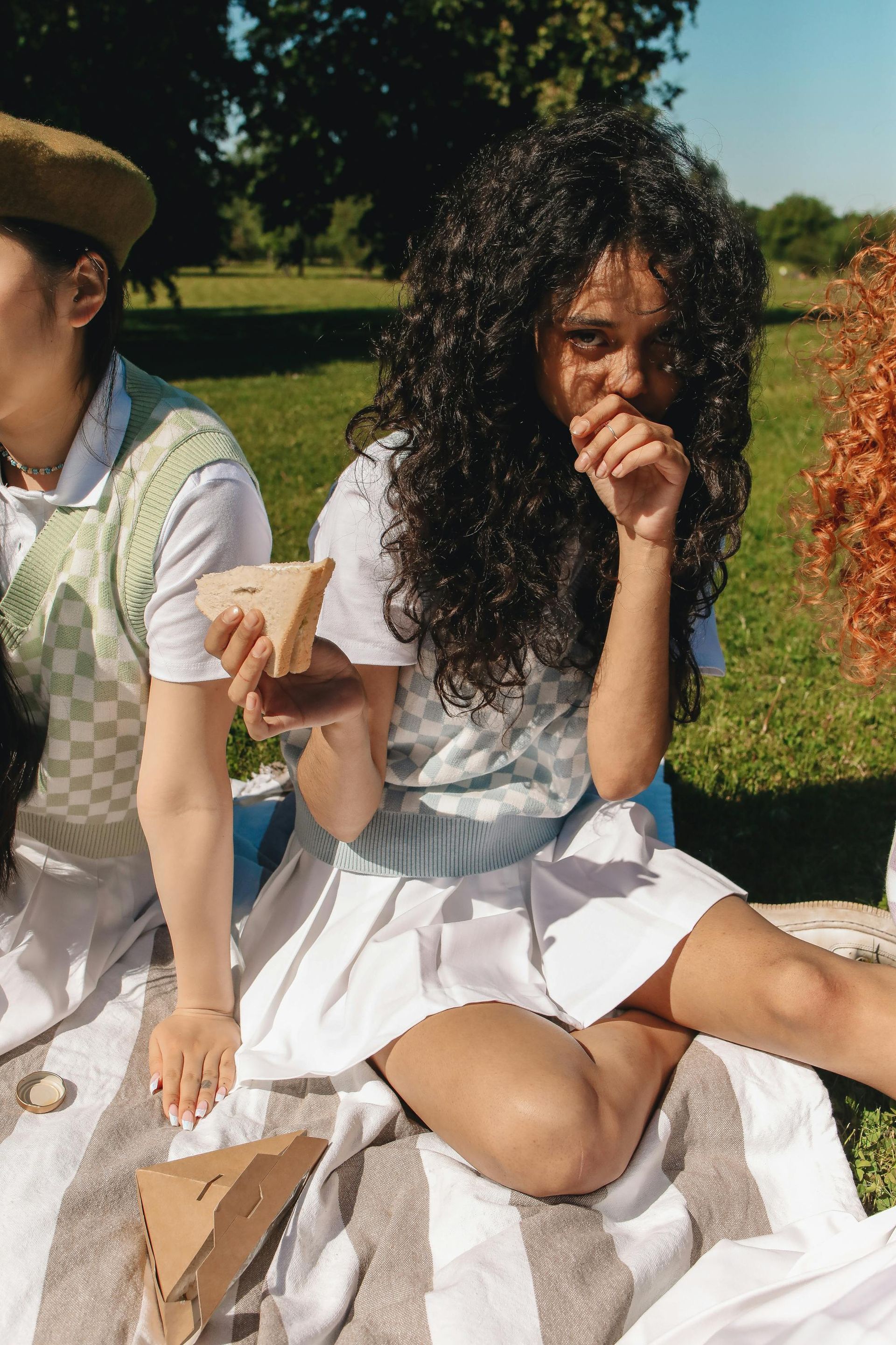 Woman eating a sandwich while sitting on a picnic blanket in a park. Wearing white skirt and vest.