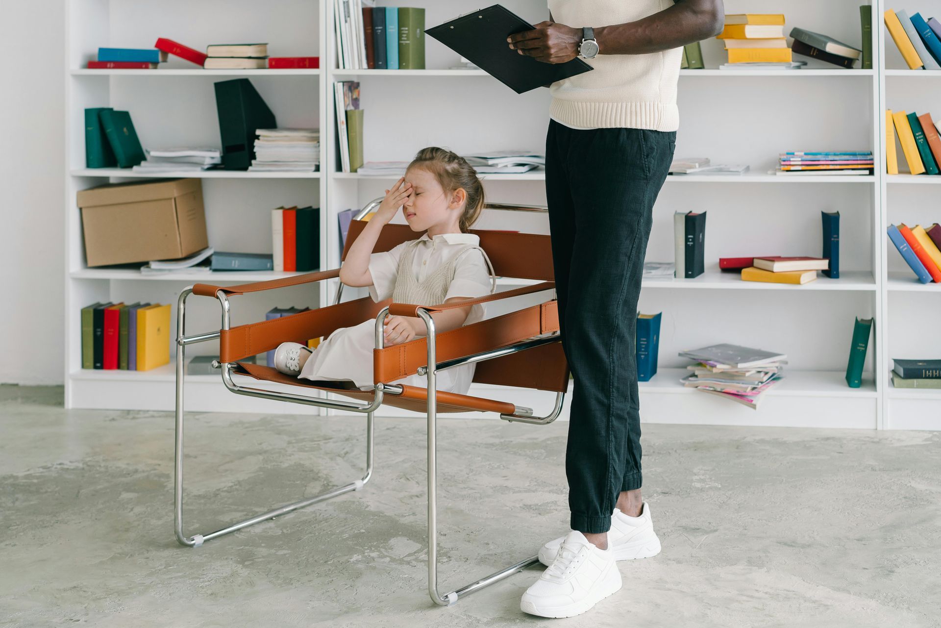 Child sits in a leather chair, looking thoughtful, while an adult stands holding a clipboard near a bookshelf.