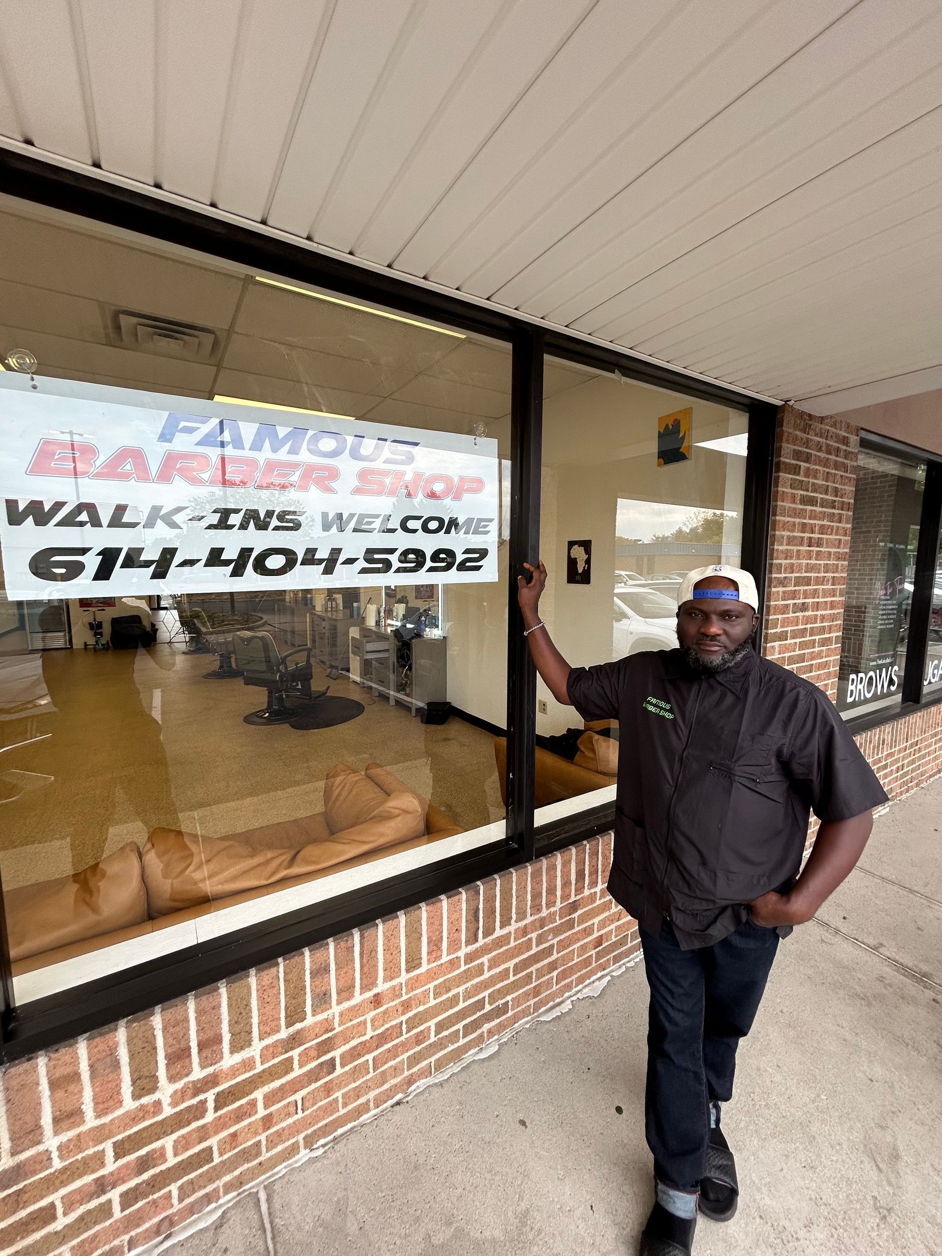 Barber standing outside Famous Barber Shop with a window sign displaying the shop's name and phone number.