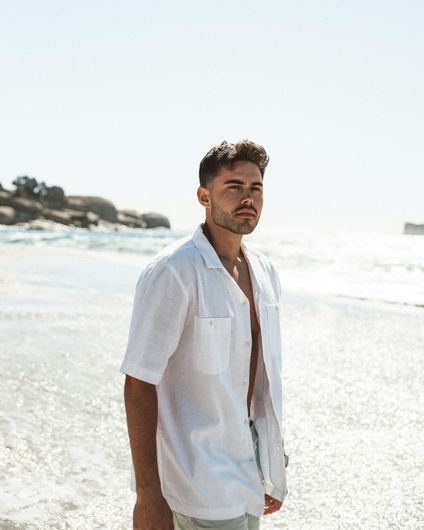 Man in a white shirt standing on a sunny beach with waves in the background.