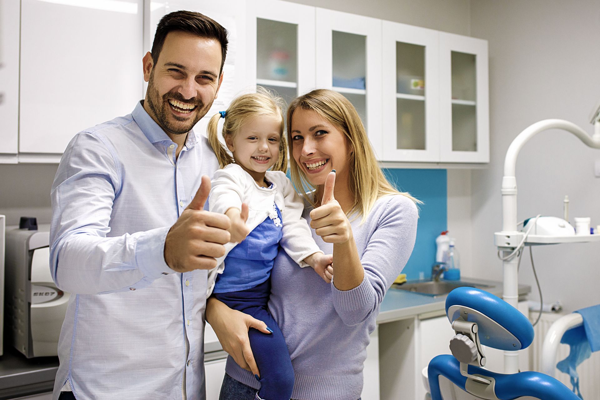 Two adults and a child giving thumbs up inside a dental clinic, with a dental chair visible. Two adults and a child giving thumbs up inside a dental clinic, with a dental chair visible.