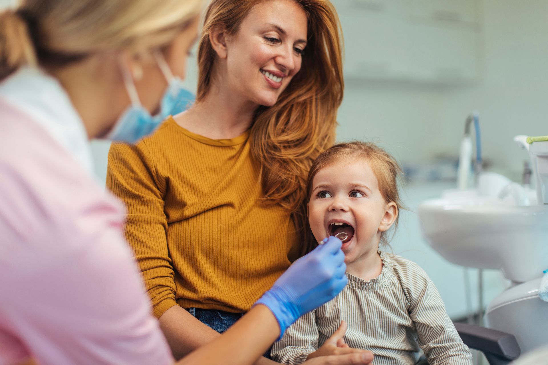 Dentist examines young child seated on parent's lap in a friendly dental clinic setting.