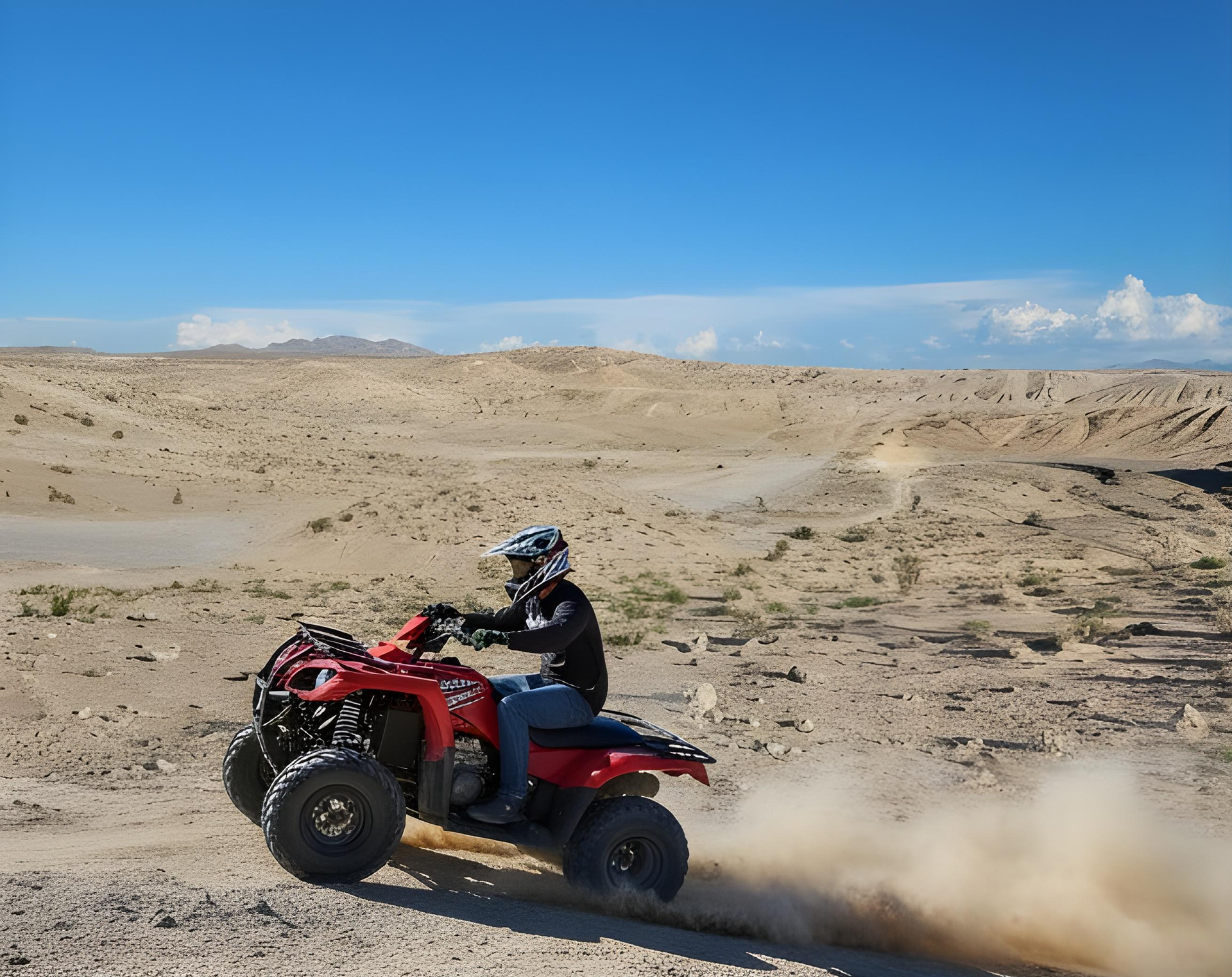 A person wearing safety gear rides a red ATV through a dusty, arid desert landscape under a clear blue sky.