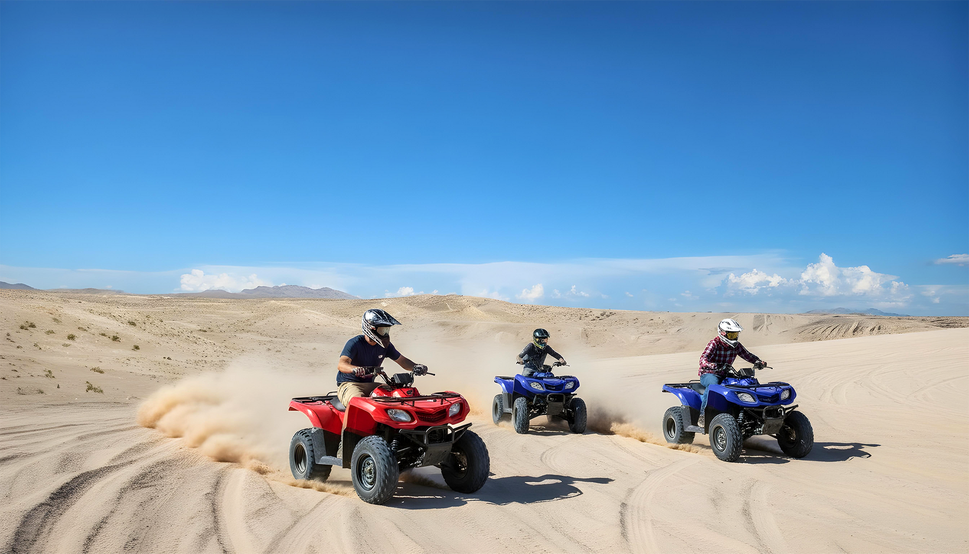 Three people ride ATVs across a sunny, sandy desert landscape, kicking up dust as they drive.