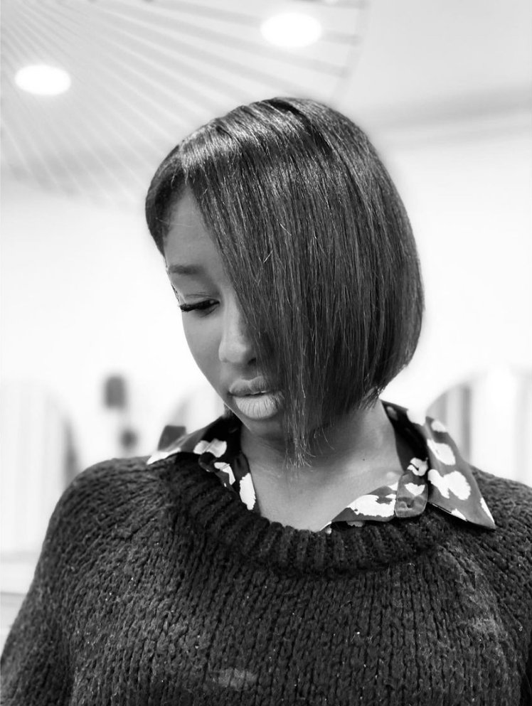 Femme aux cheveux coupés au carré raides et anguleux, regardant vers le bas. Elle porte un pull et une chemise à col.