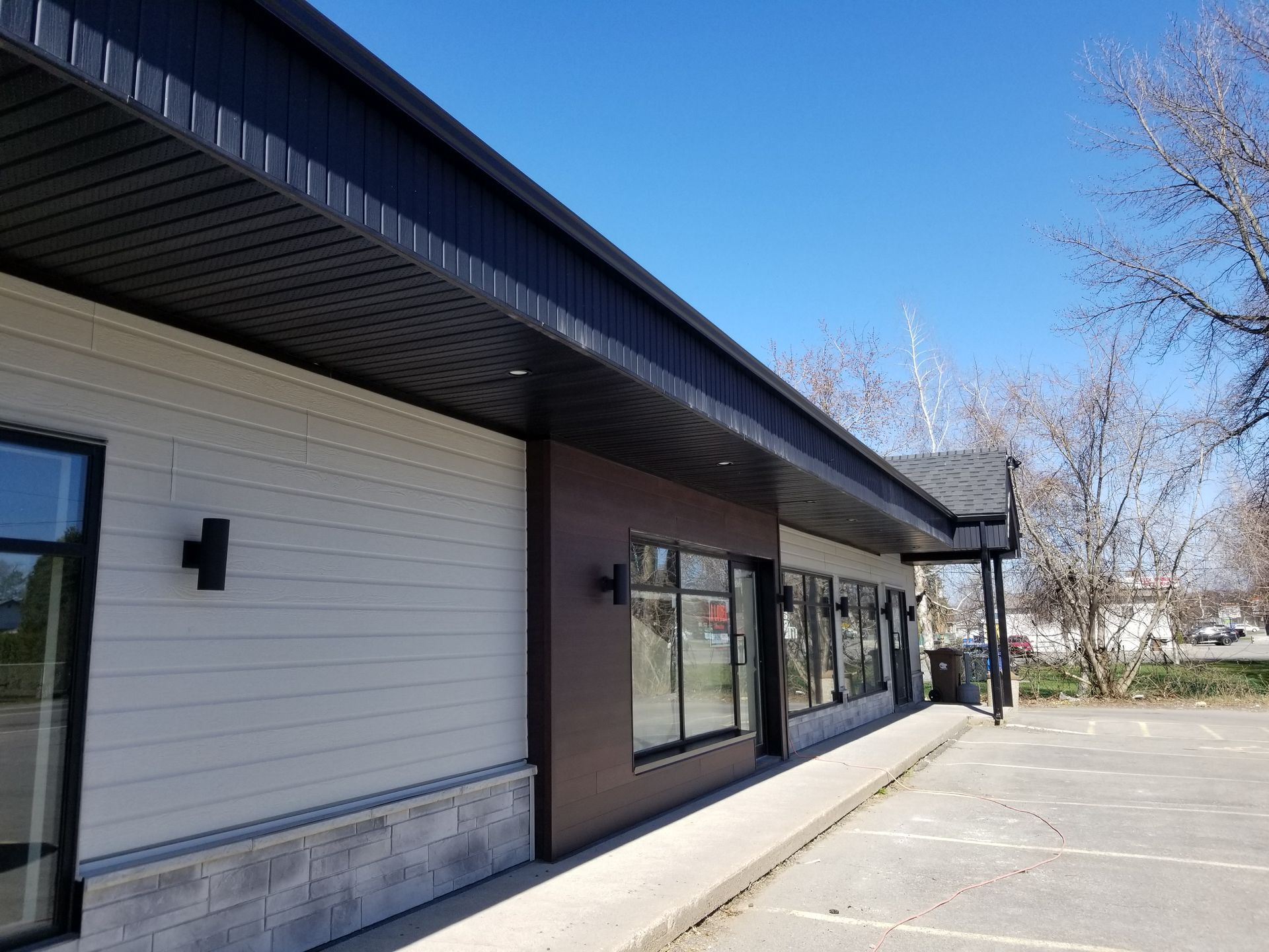 Vue en angle de l'extérieur d'un bâtiment commercial avec un bardage horizontal clair, des panneaux bruns et des fondations en pierre.