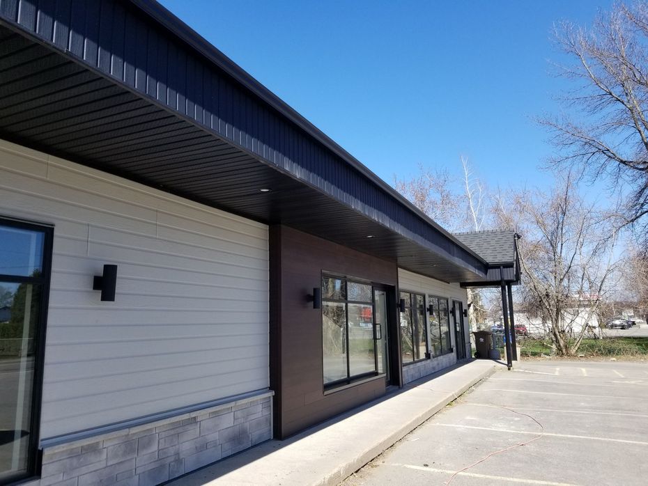 Vue en angle de l'extérieur d'un bâtiment commercial avec un bardage horizontal clair, des panneaux bruns et des fondations en pierre.