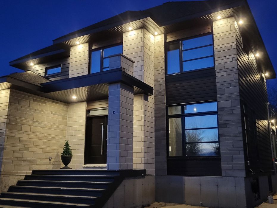 Extérieur d'une maison moderne à deux étages au crépuscule, avec des accents de pierre claire, des lambris foncés, un éclairage encastré et des marches d'entrée.