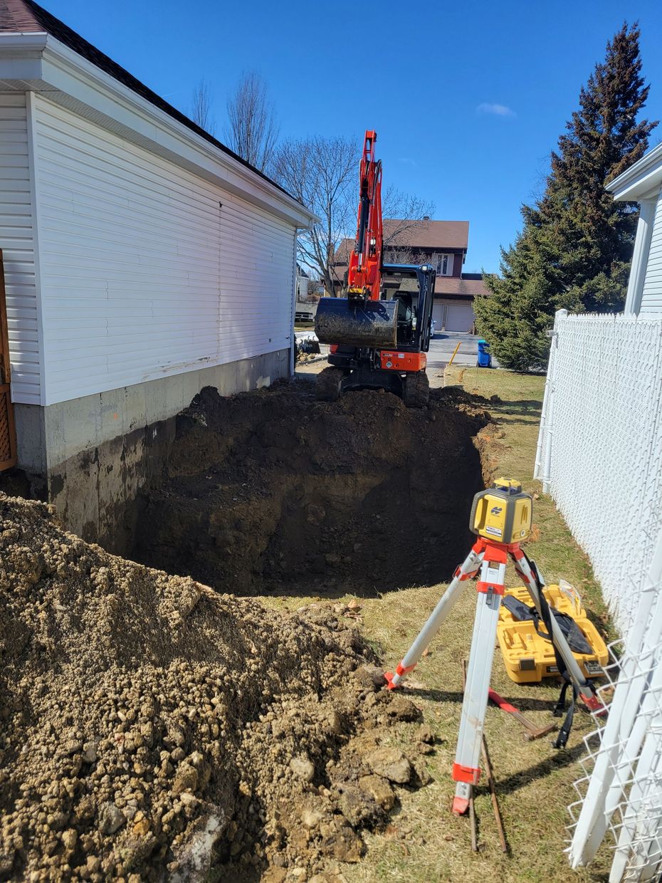 Une pelleteuse rouge creuse une tranchée le long des fondations d'une maison blanche, avec un niveau laser sur un trépied au premier plan.