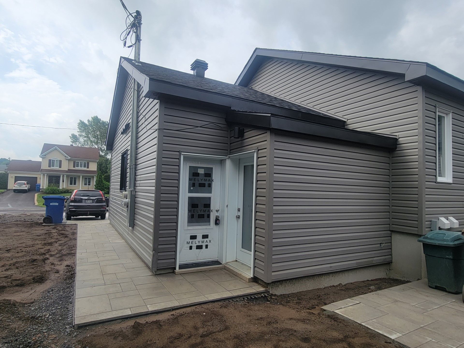 Vue extérieure d'une maison aux murs gris, avec deux portes blanches, une allée pavée et une poubelle verte, par temps nuageux.