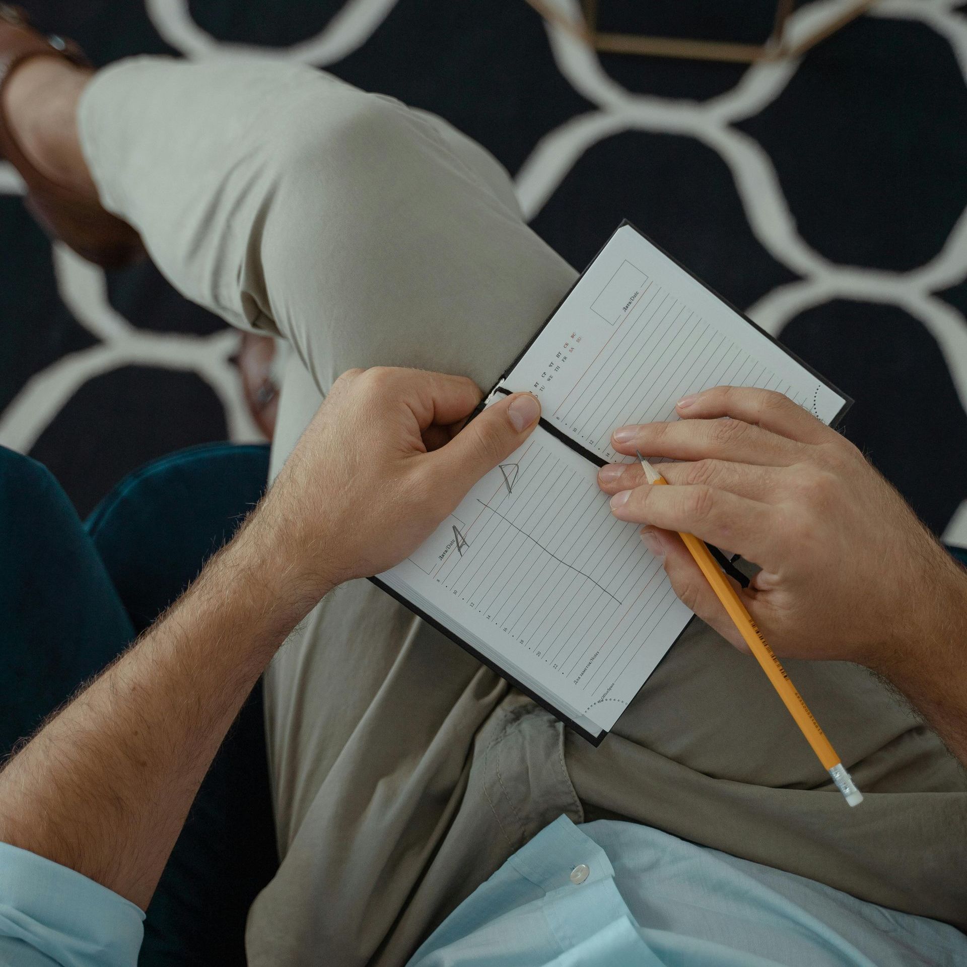 A man is sitting on a couch writing in a notebook with a pencil.