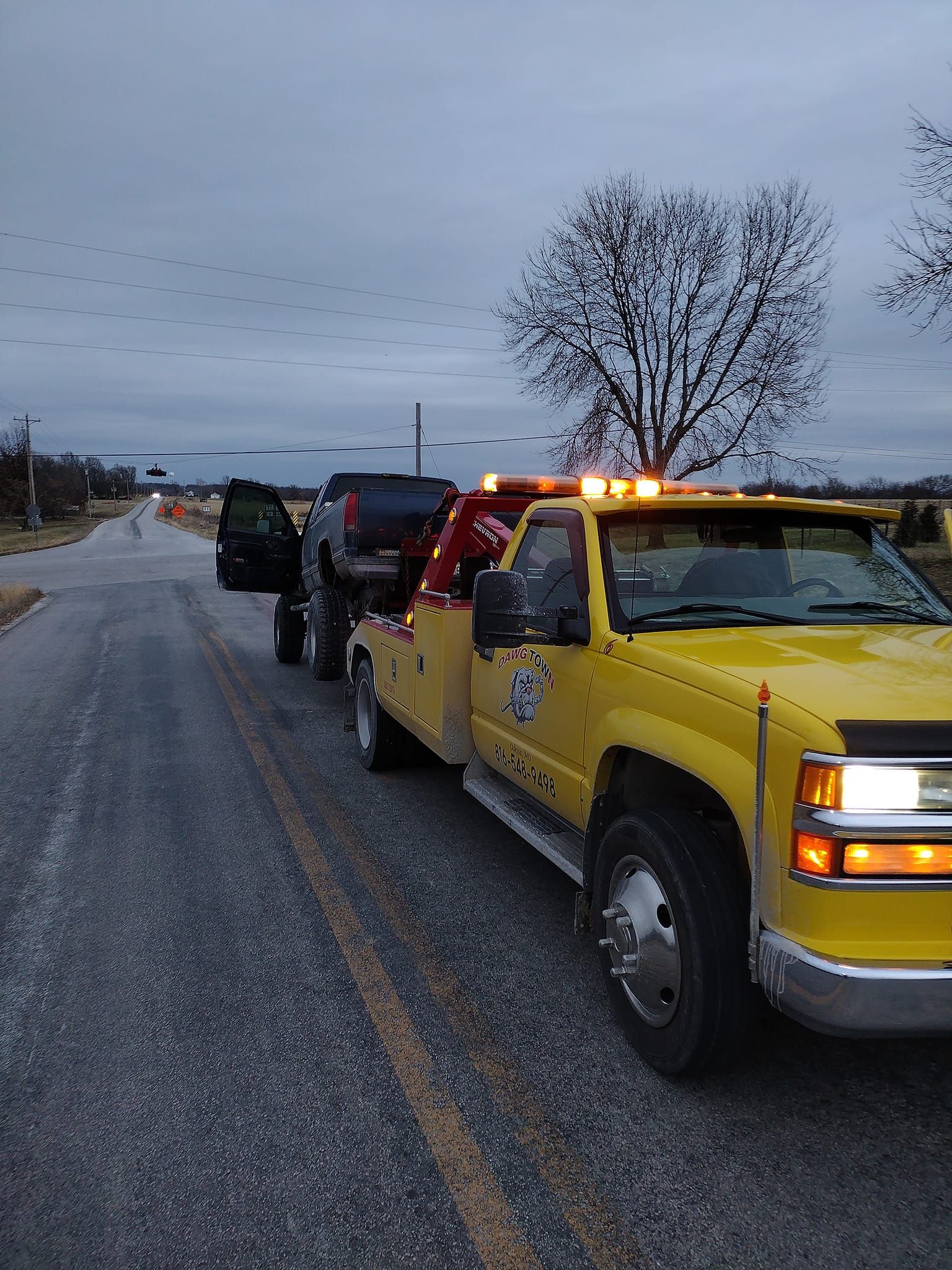 A yellow tow truck is towing a black truck down a road.