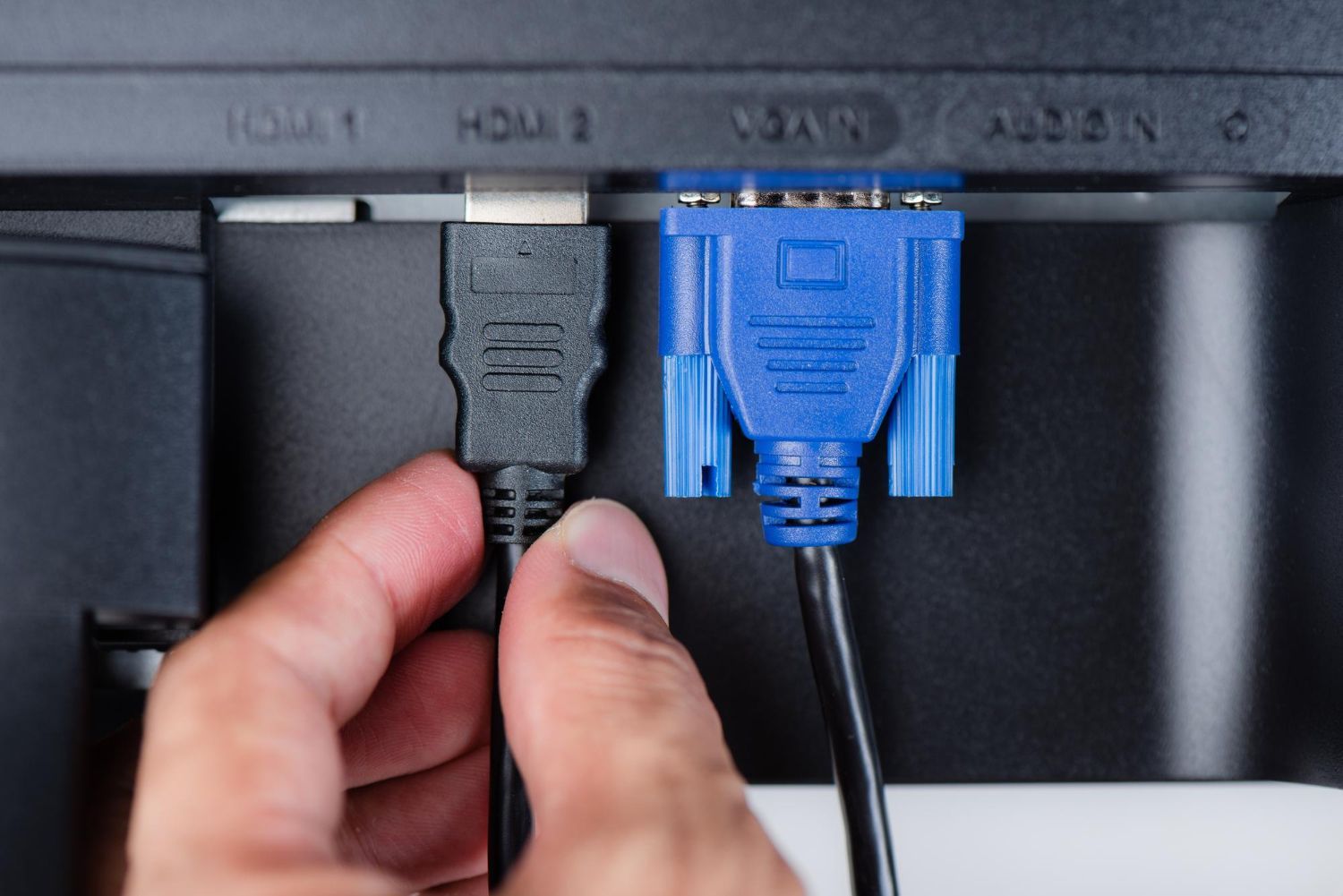 A hand plugging a black HDMI cable into a port next to a blue VGA cable on the back of a monitor.