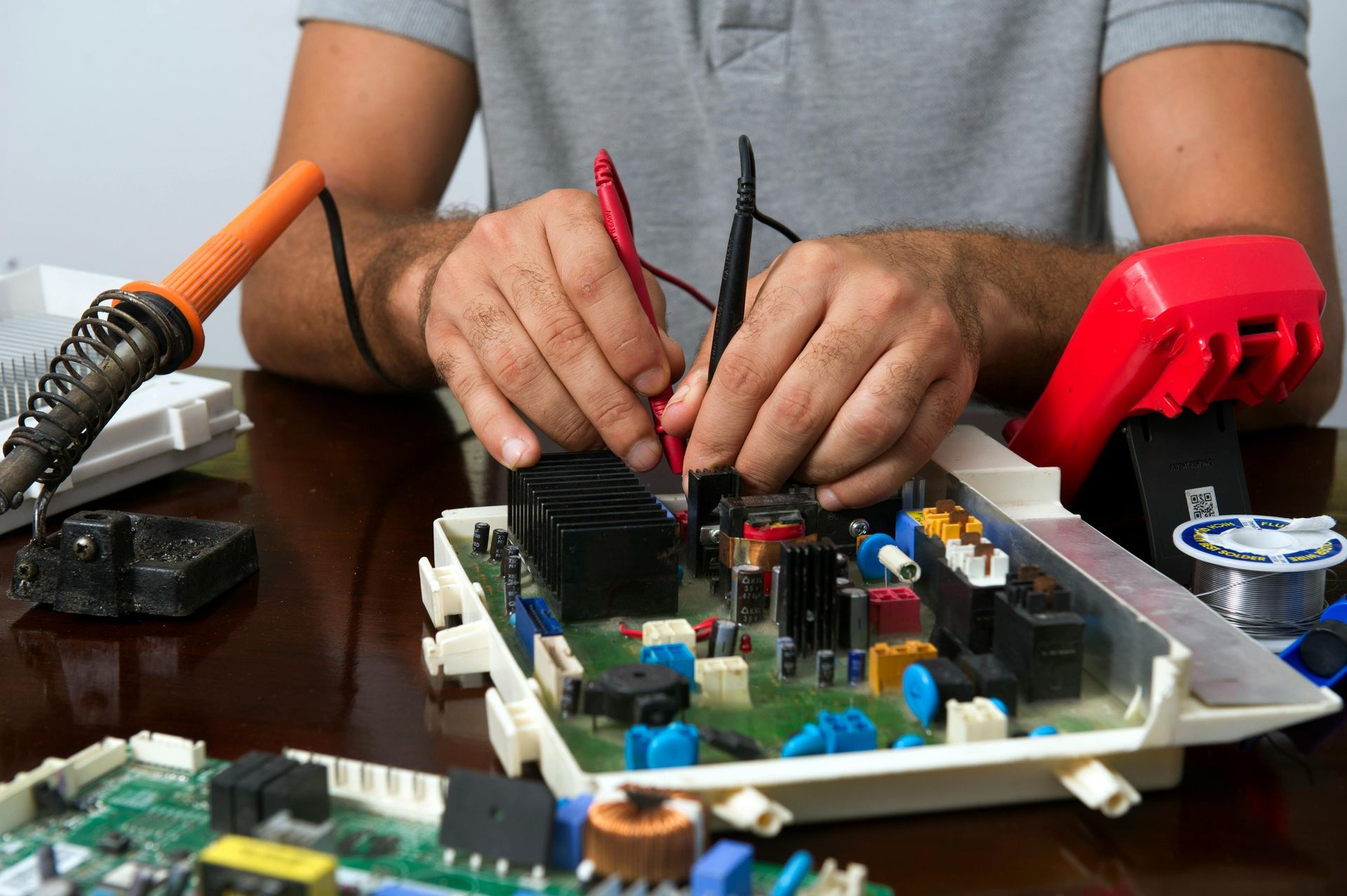 A person repairing an electronic circuit board with a multimeter and a soldering iron on a wooden table.