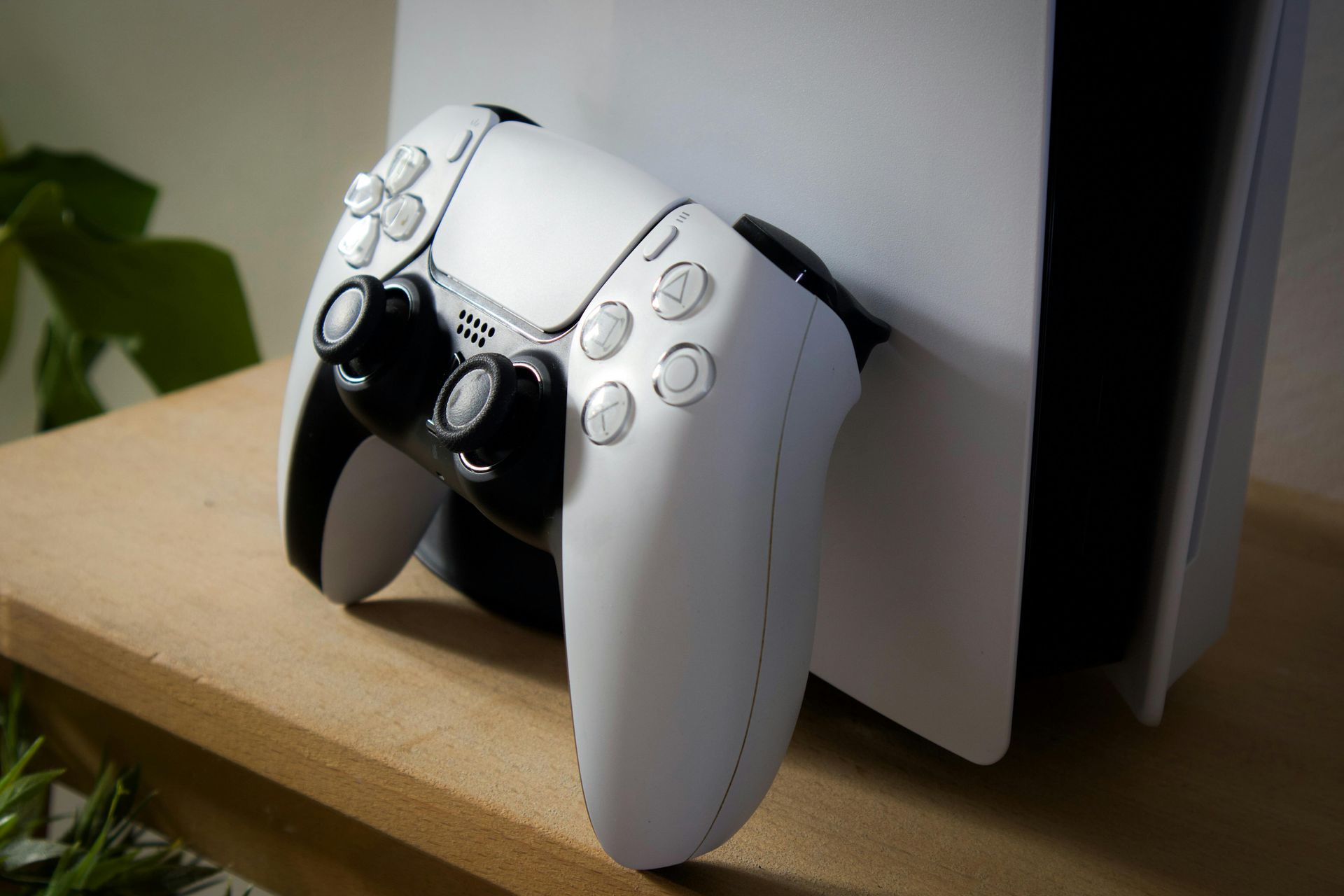 A white PlayStation 5 DualSense controller leaning against a white PlayStation 5 console on a wooden surface.