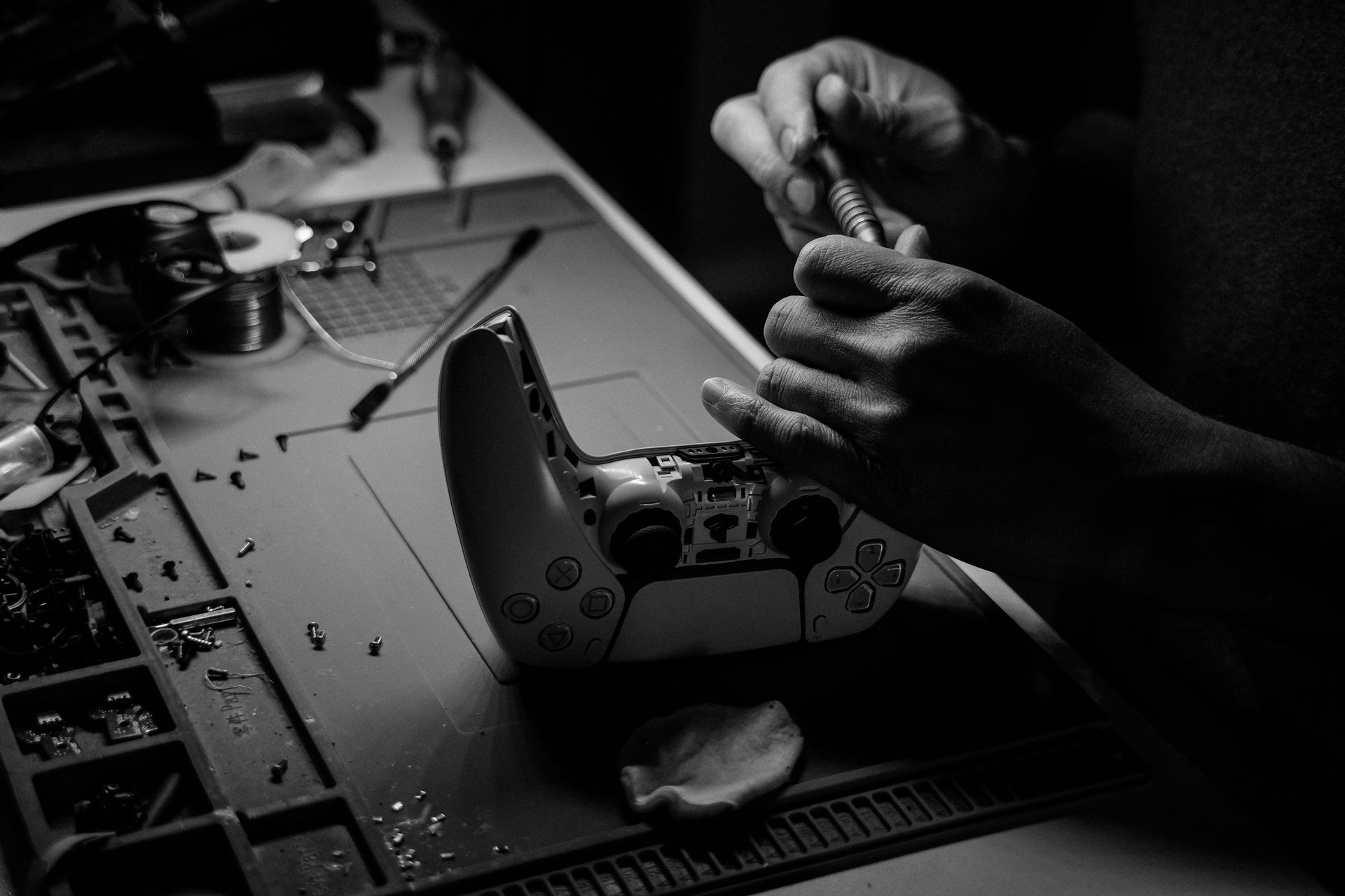 A black-and-white close-up of a person using a precision screwdriver to repair a game controller on a workbench.