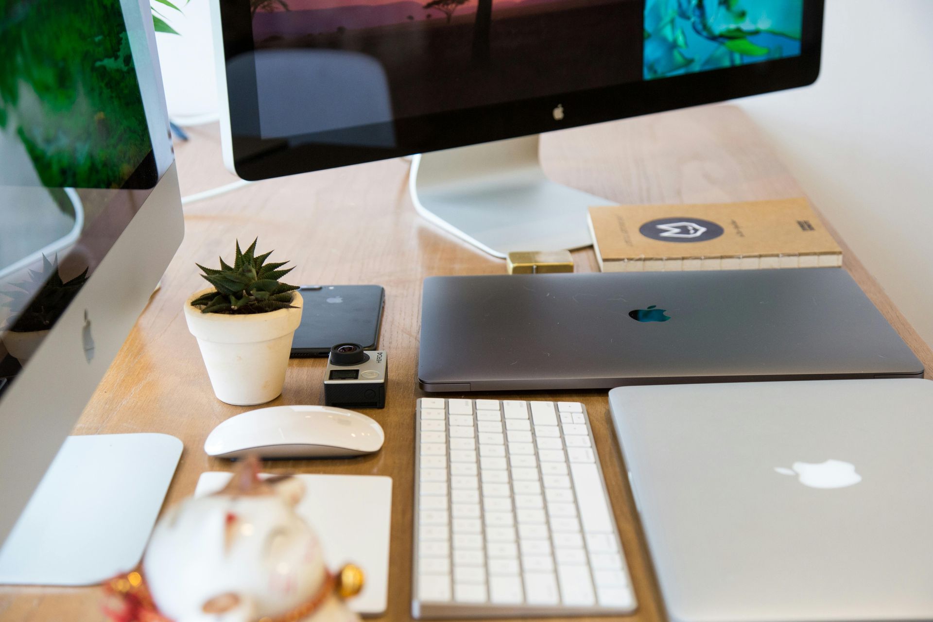 A clean desk setup featuring two computer monitors, a closed MacBook, a keyboard, a mouse, a plant, and a notebook.