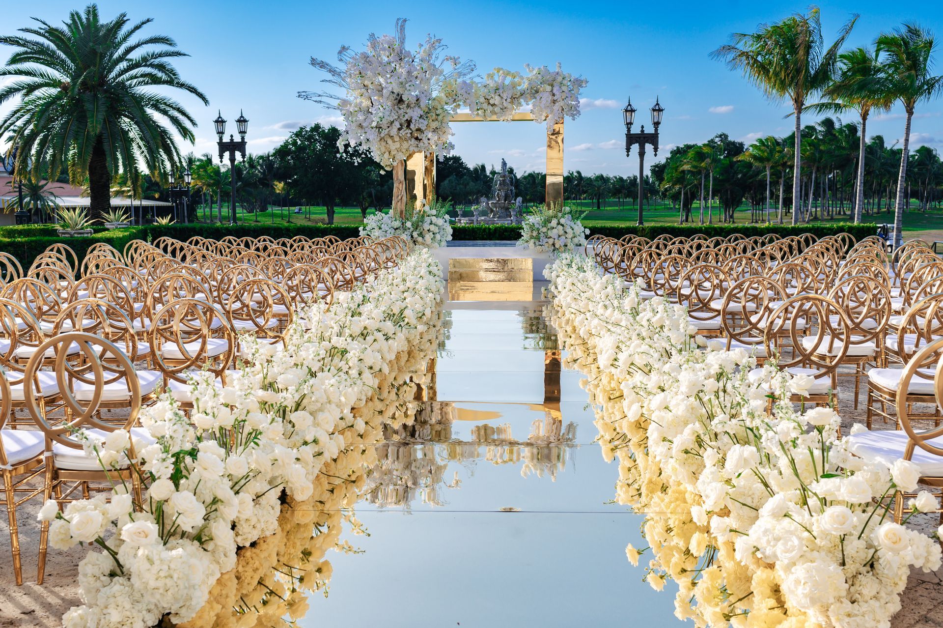 a wedding ceremony is taking place in a park with a long aisle lined with white flowers and chairs .
