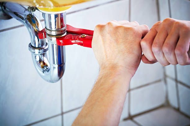 Hands using a red wrench to tighten a chrome p-trap pipe under a sink against a tiled wall.
