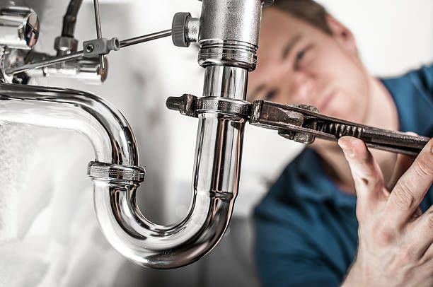 A person in a blue shirt uses a wrench to tighten a charome pipe connection under a sink.