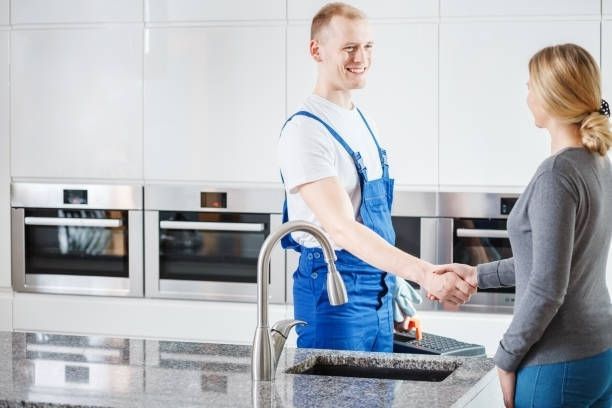 A technician in blue overalls shakes hands with a customer in a modern kitchen with stainless steel appliances.