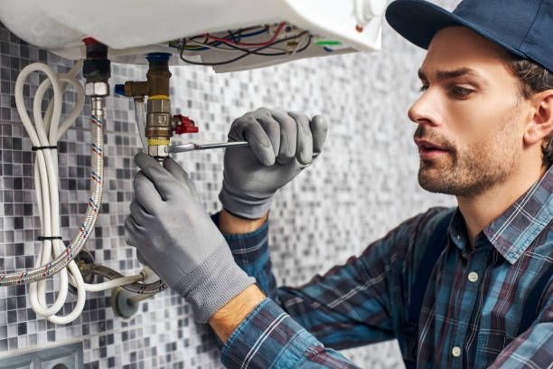 A person wearing work gloves and a cap uses a wrench to adjust a pipe valve on a wall-mounted water heater.